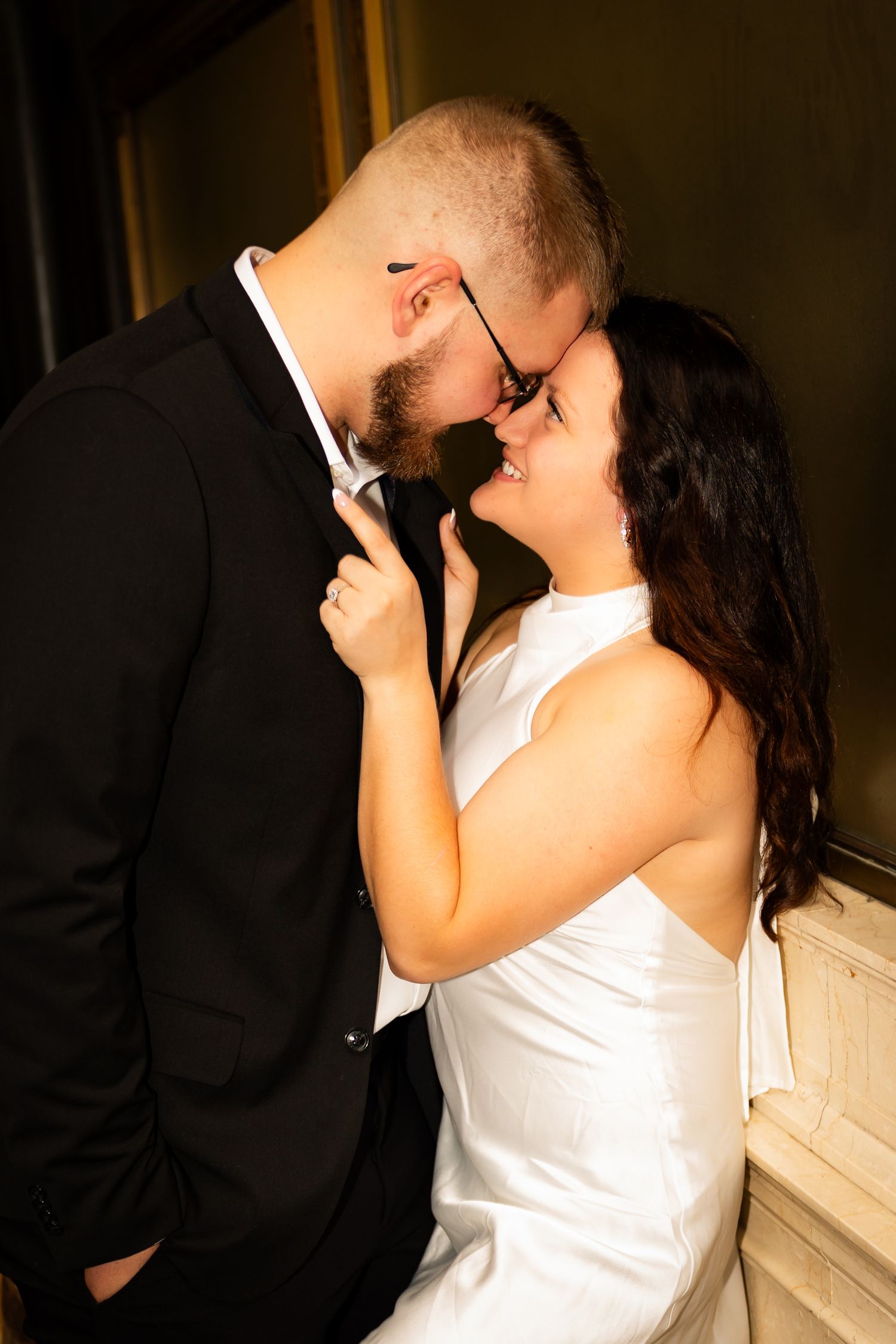 Intimate moment shared between couple in formal attire as they lean in close together against light colored wall.
