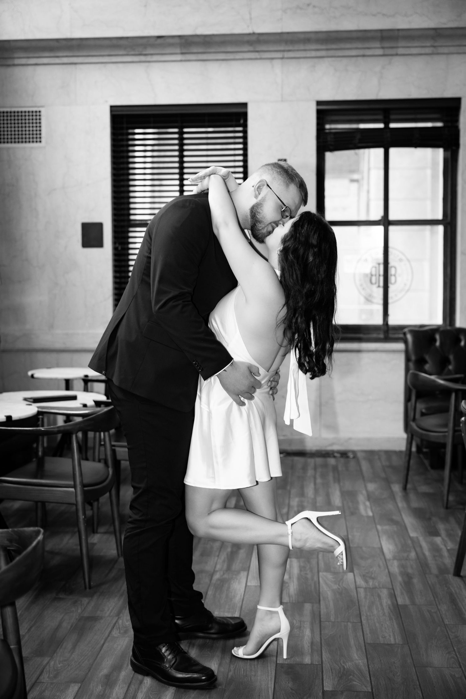 A romantic couple in formal attire sharing a passionate kiss while she leans back, her high heels lifted off the hardwood floor.