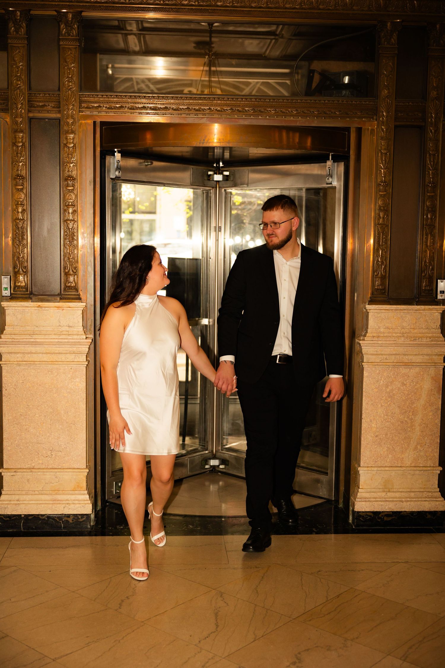 A couple in formal attire stands by an elegant elevator entrance with ornate gold trim details in a luxurious building.
