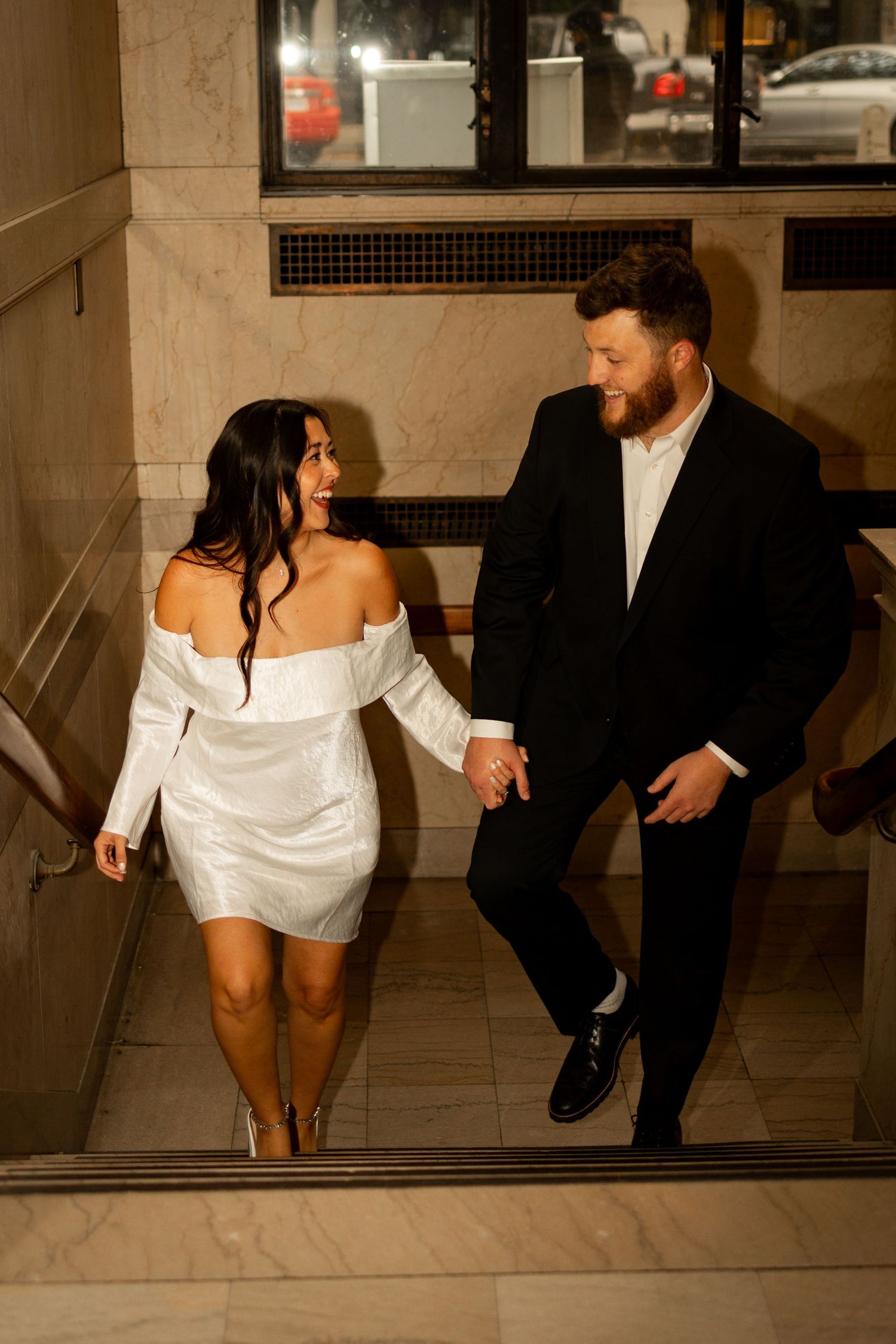 A couple walks down marble stairs, with her wearing a white off-shoulder dress and him in a black suit at night.