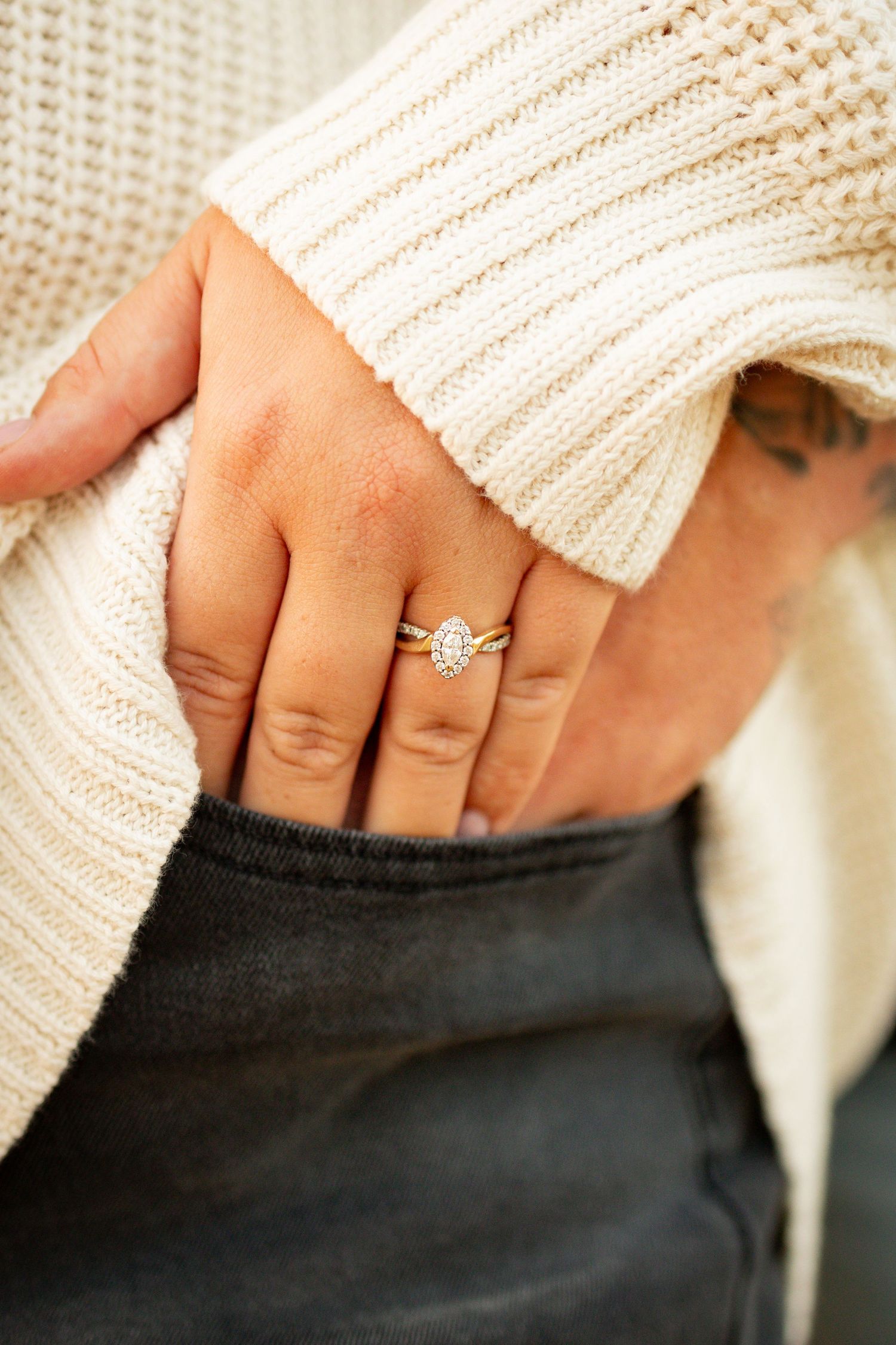 Close up of a sparkling oval diamond engagement ring on hand wearing cream knit sweater and dark pants.