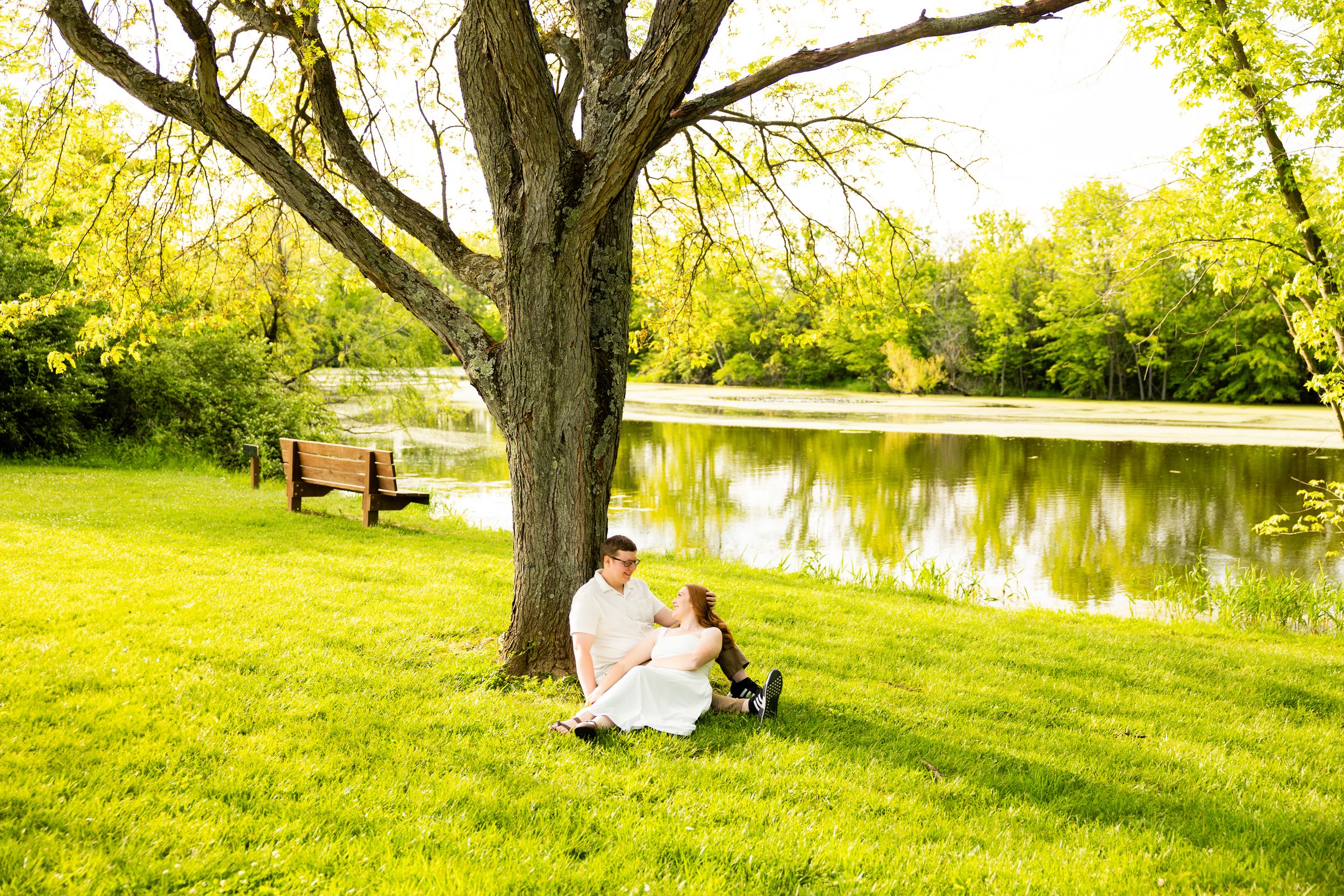 Couple sitting together under a large tree by a peaceful pond on lush green grass in a sunny park.
