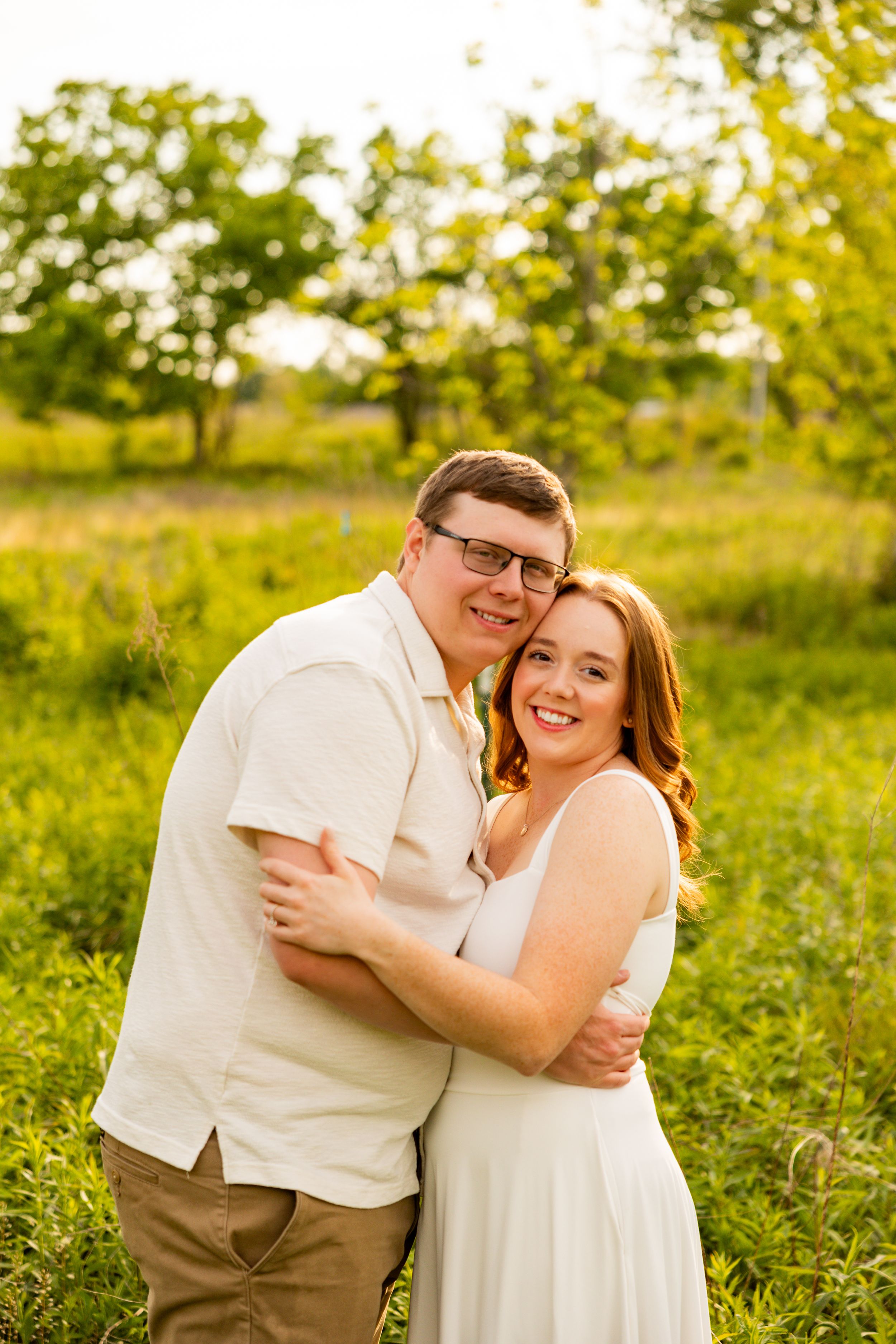 A smiling couple embraces outdoors in a lush green field during golden hour, wearing coordinating white and tan outfits.