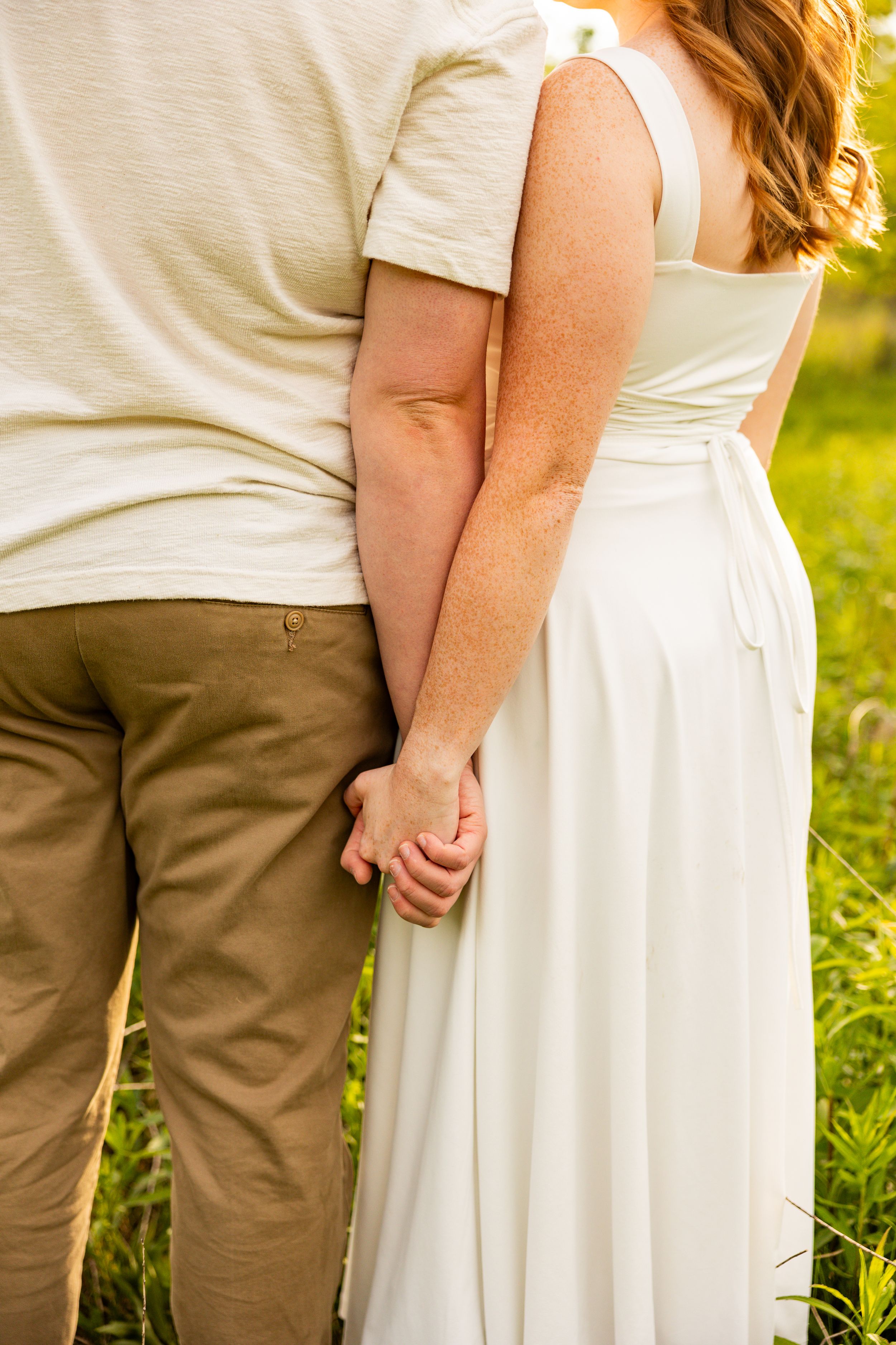 Couple holding hands from behind, woman in white dress and man in khaki pants, standing in a sunny green field.