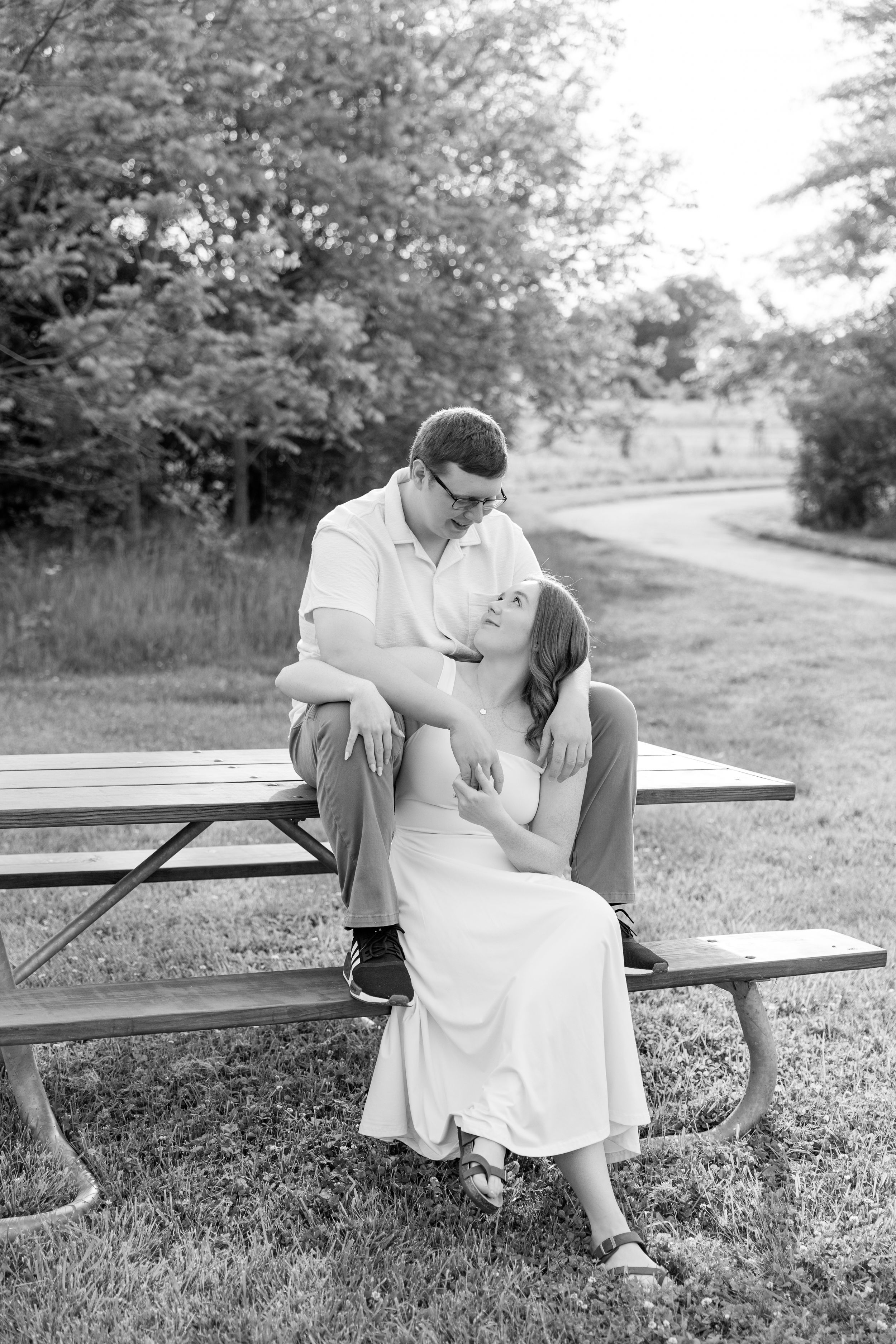 Black and white photo of a couple sitting together at a picnic table in a park, sharing a tender romantic moment.