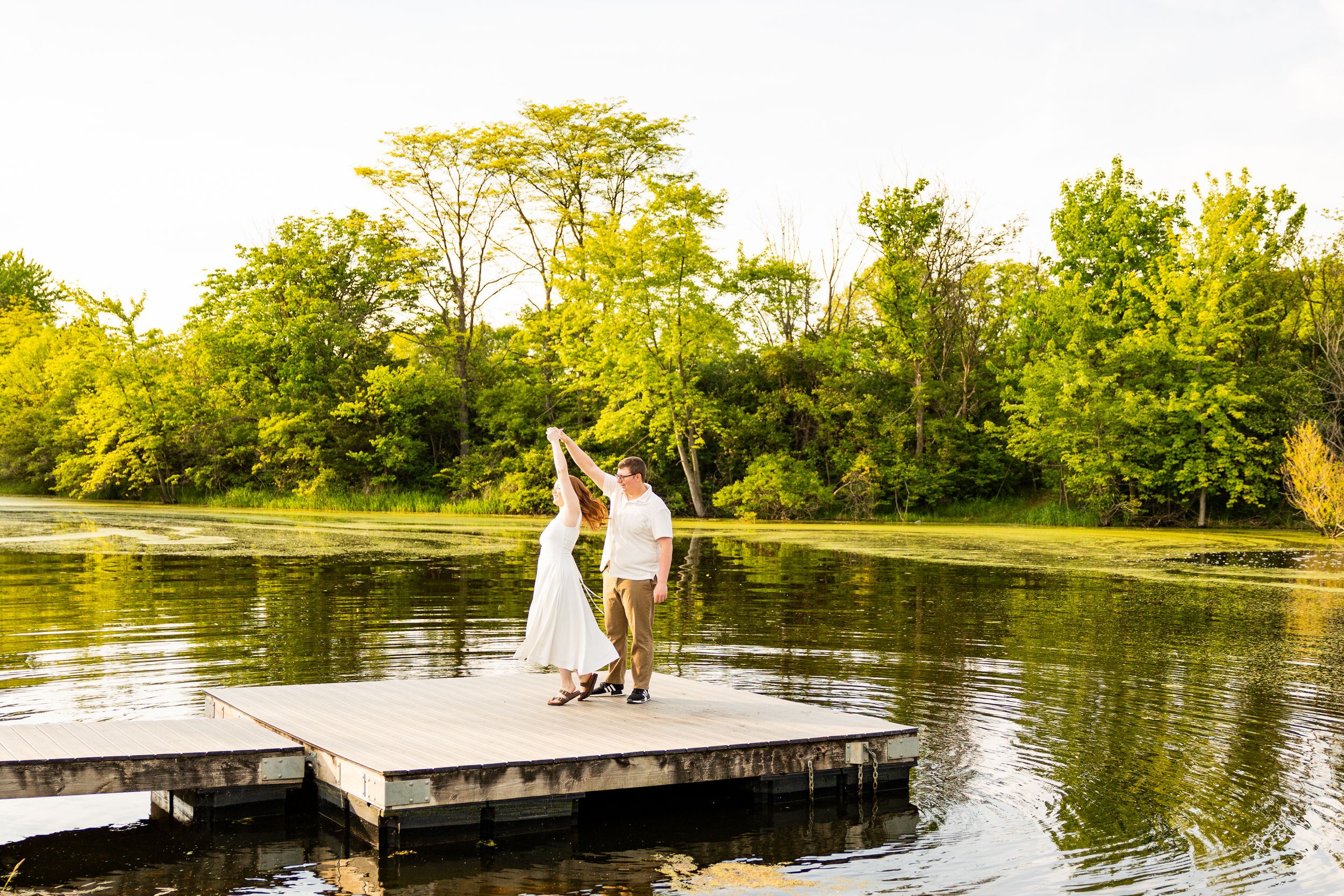Couple in white attire dancing on a lakeside dock surrounded by lush green trees during golden hour.