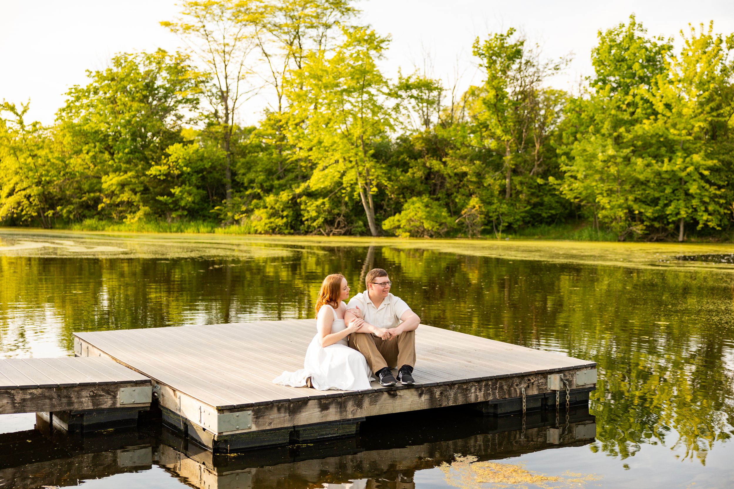 Couple sitting together on a wooden dock over a calm lake surrounded by lush green trees at sunset.