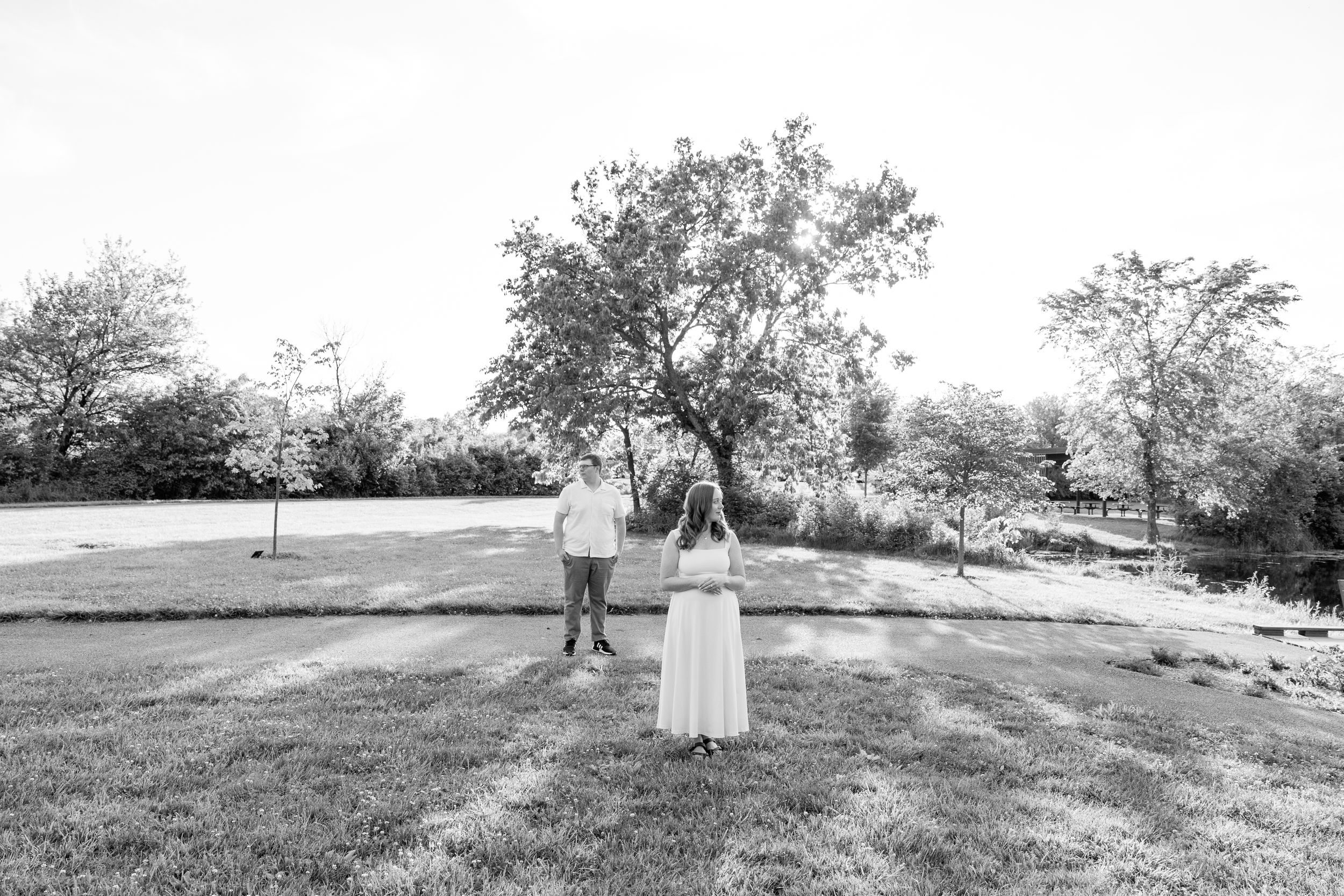 Black and white photo of a couple standing apart on a grassy lawn with trees in the background.