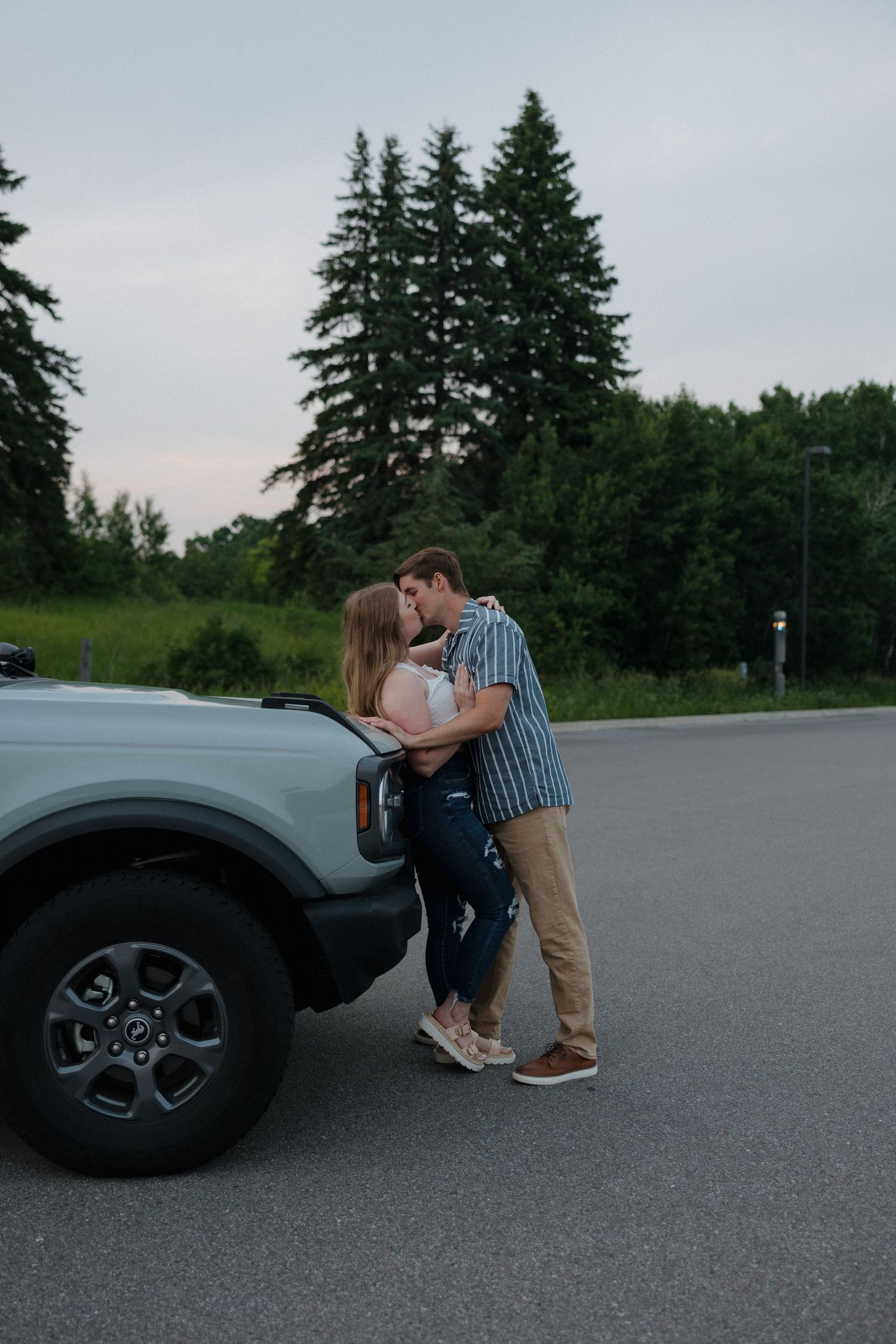 A couple shares a romantic moment leaning against a vehicle in a parking area near evergreen trees at dusk.