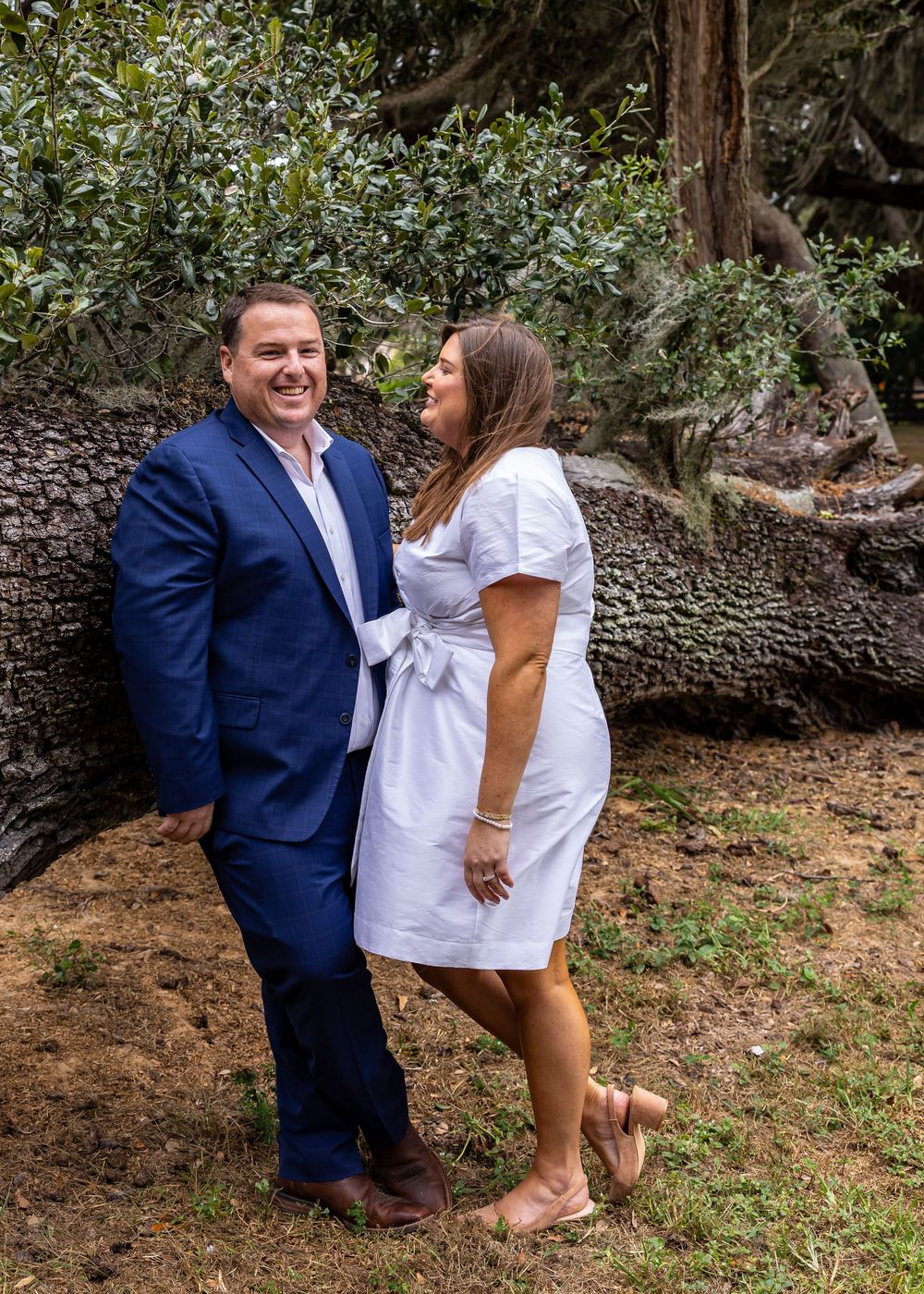 Playful couple poses together under the branches of a majestic oak tree.