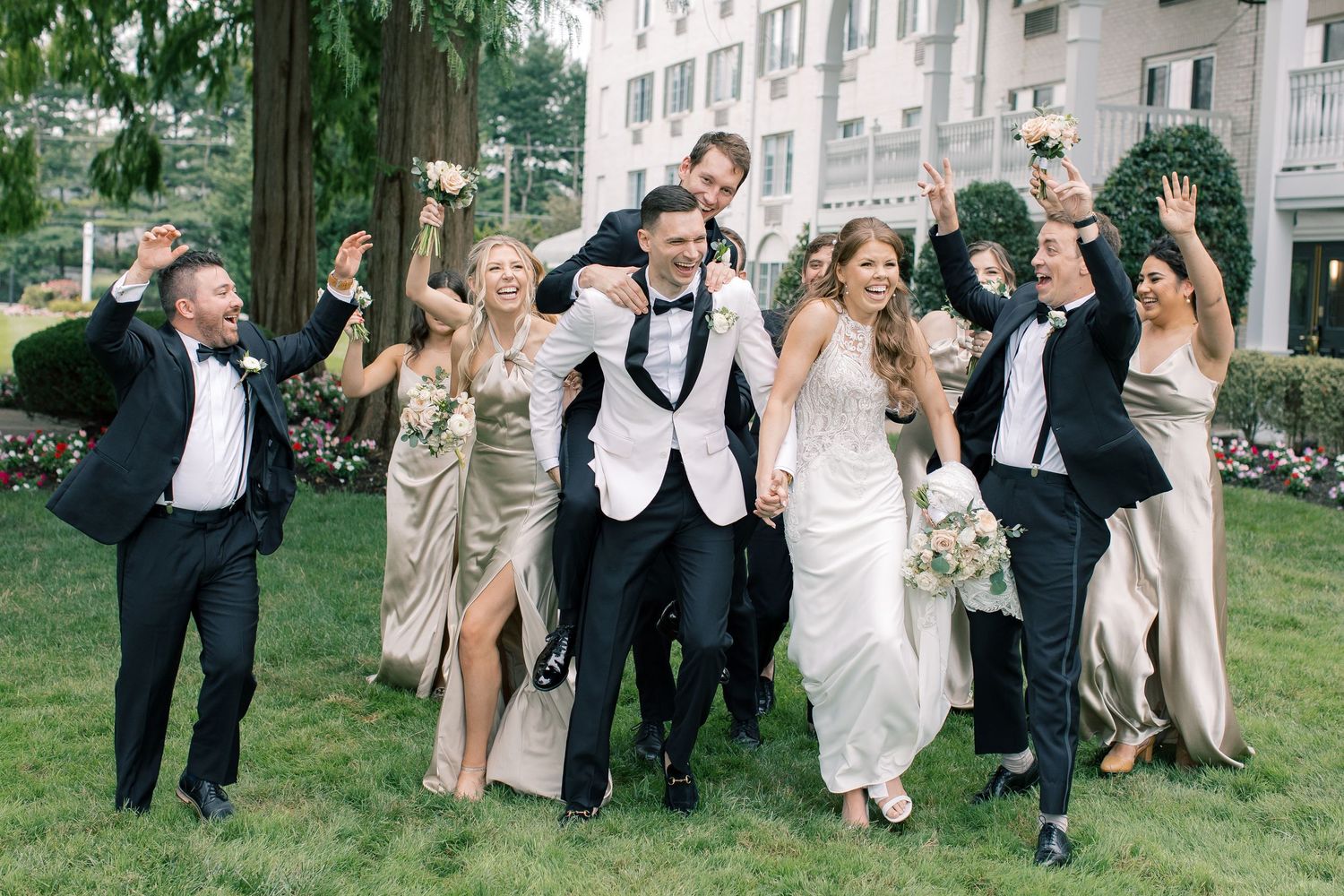 Wedding party jumps joyfully on grass lawn with elegant outfits and formal attire during outdoor celebration.