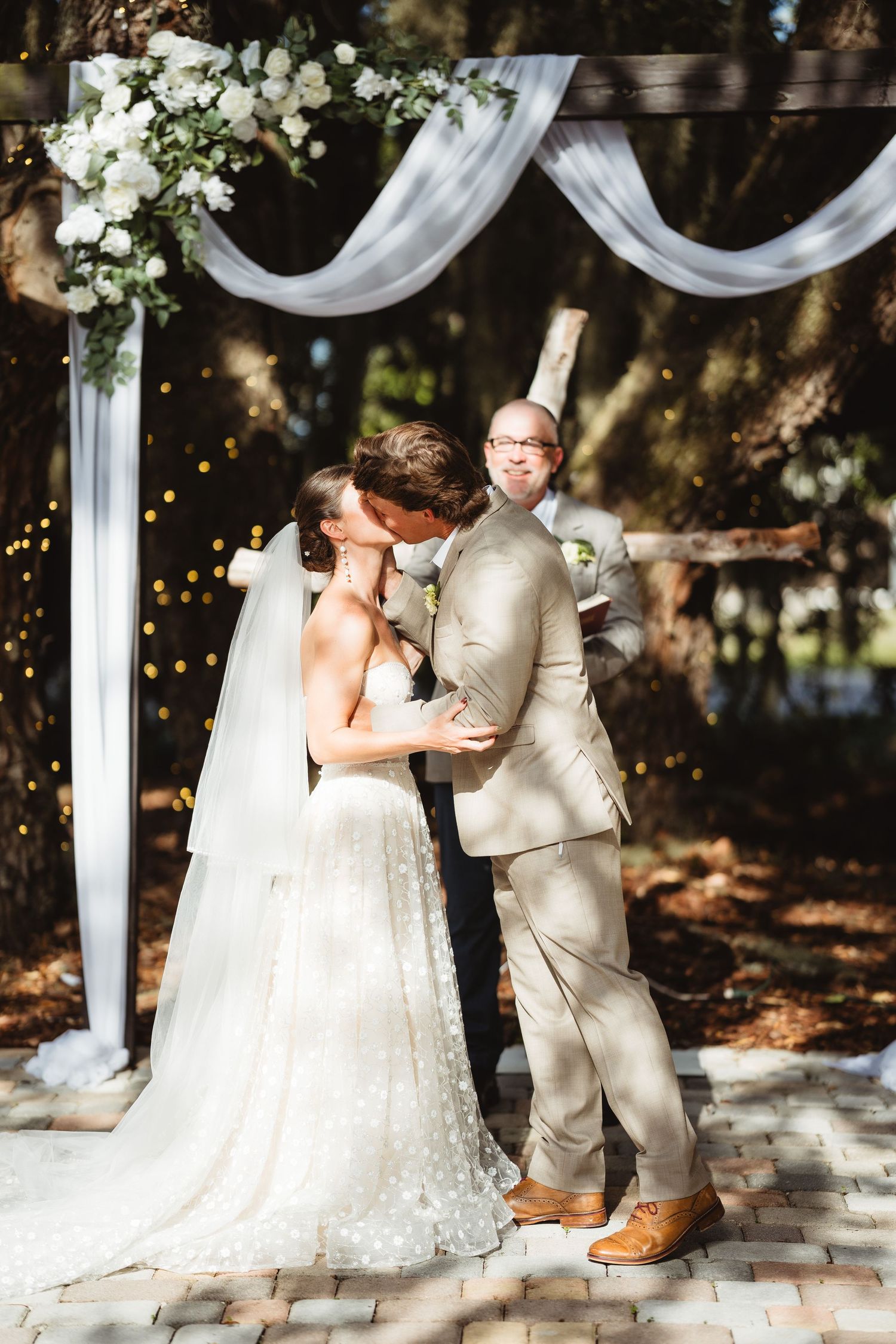 Two people share an intimate moment under an elegant white draped wedding arch decorated with white flowers in an outdoor setting.