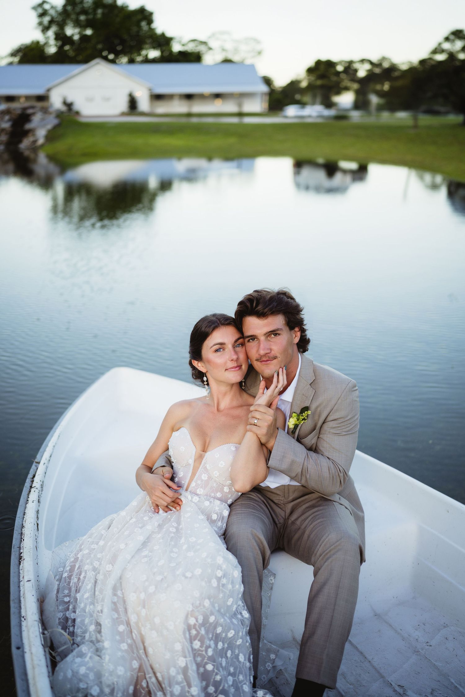 A couple shares a romantic moment in a white rowboat on a serene pond with buildings visible in the background.