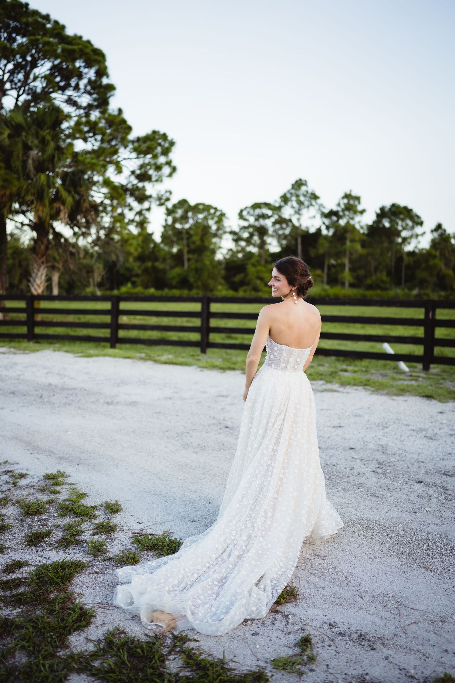 Person in flowing white lace dress walks along a gravel path with wooden fences and green trees in background.