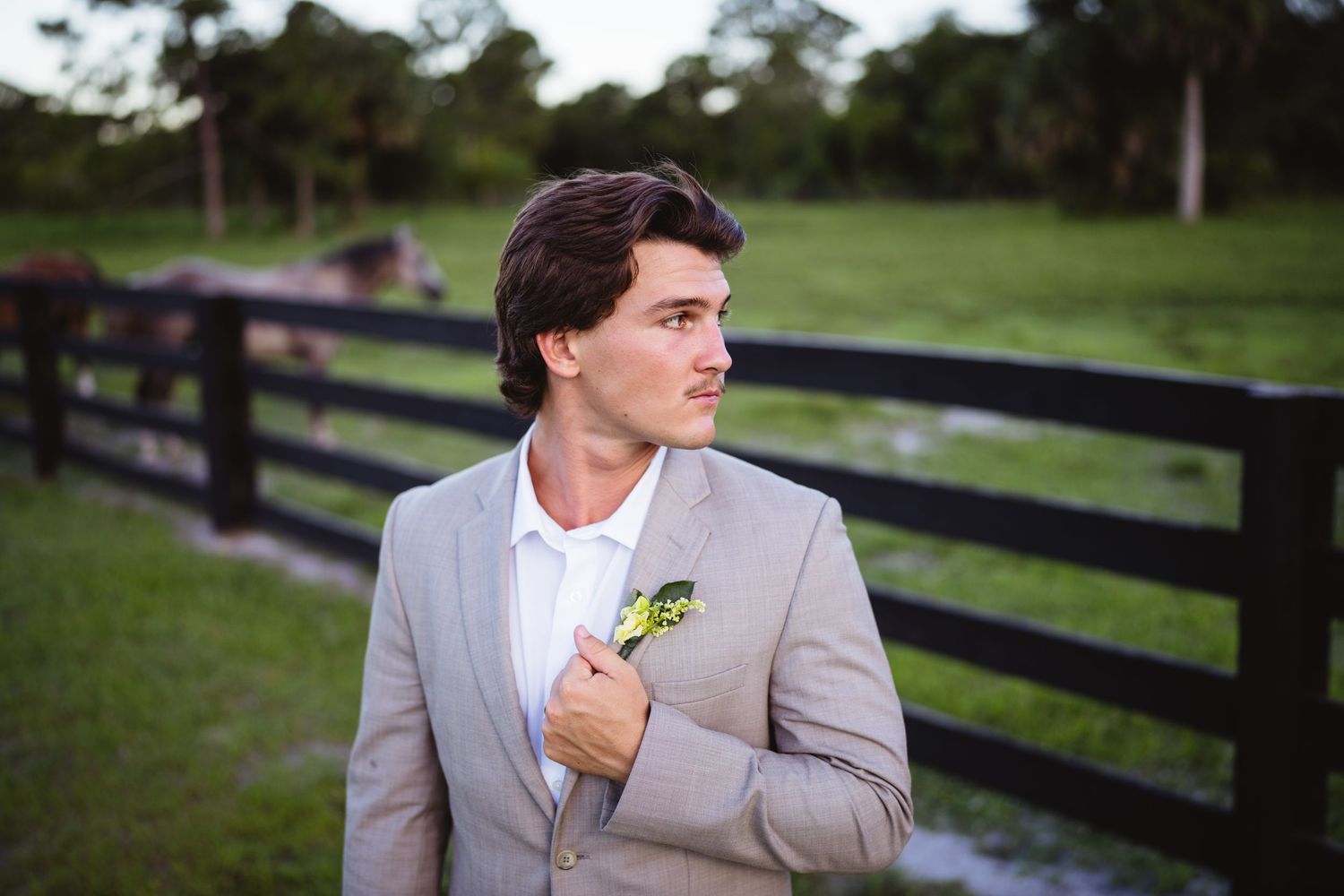 Someone in a gray suit with a boutonniere stands near a wooden fence at an outdoor venue during golden hour.