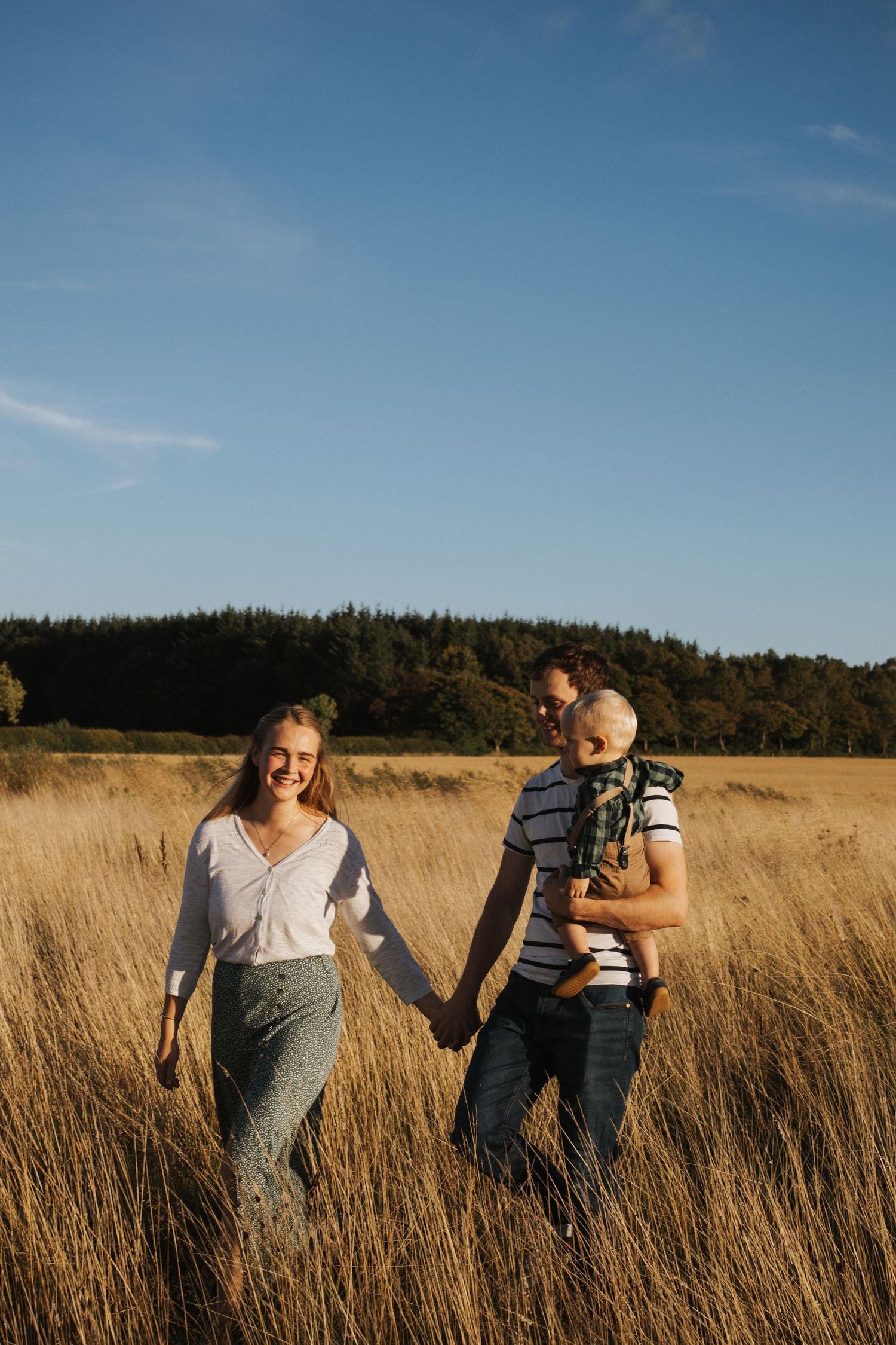 A couple holds hands while walking through a golden grass field during sunset with a forest backdrop.