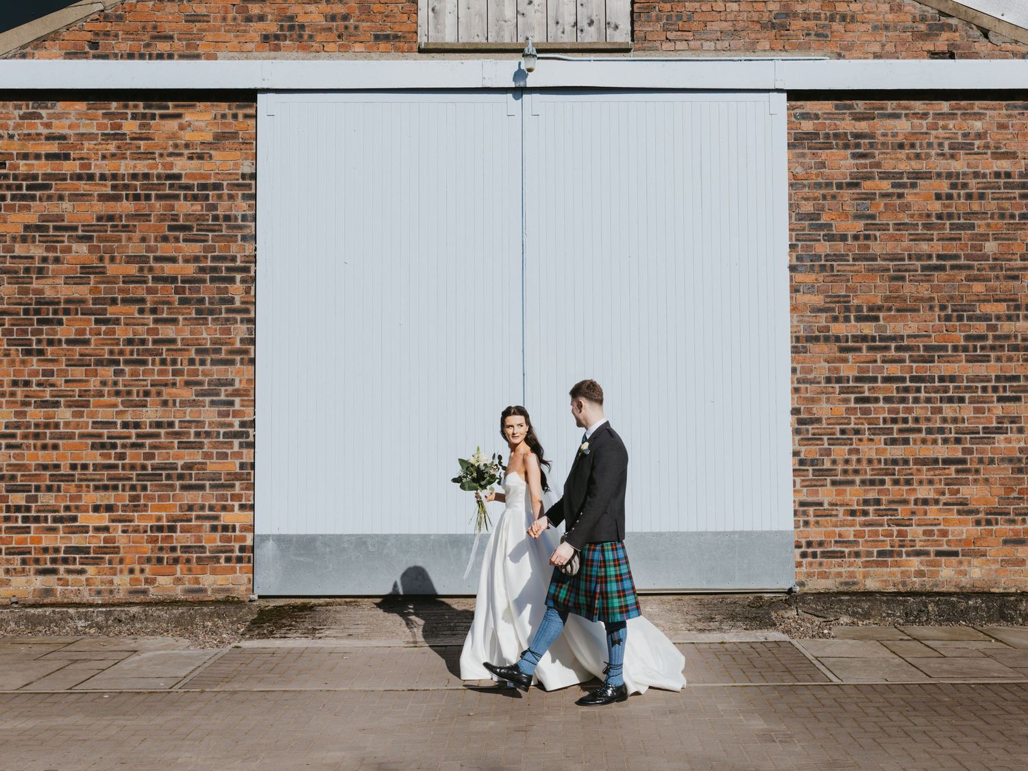 Bride and groom holding hands outside rustic brick building, groom wearing traditional Scottish tartan kilt.