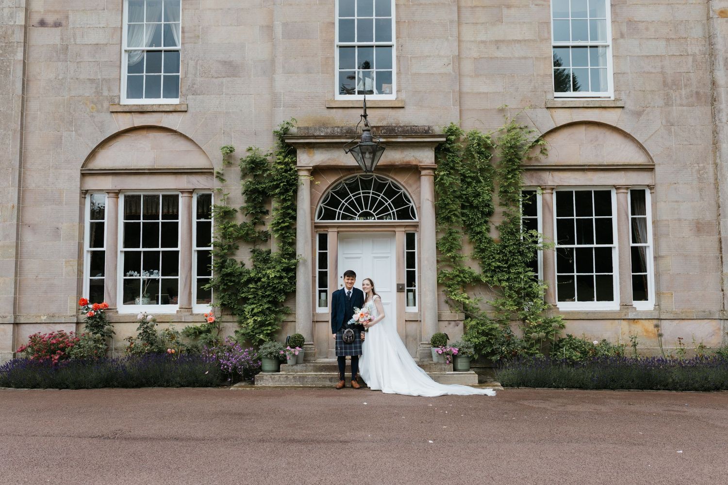 Bride and groom posing in front of an elegant stone manor house with ivy-covered walls and a classic blue door.