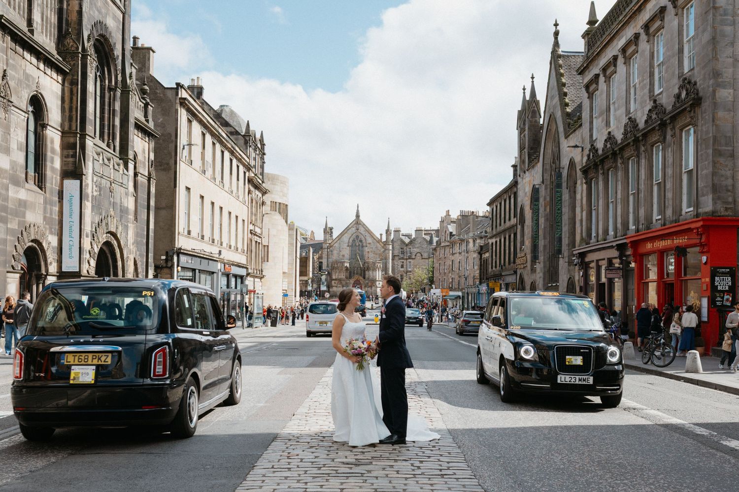 A bride and groom cross a historic cobblestone street in Edinburgh, Scotland, surrounded by classic black taxis and Georgian architecture.