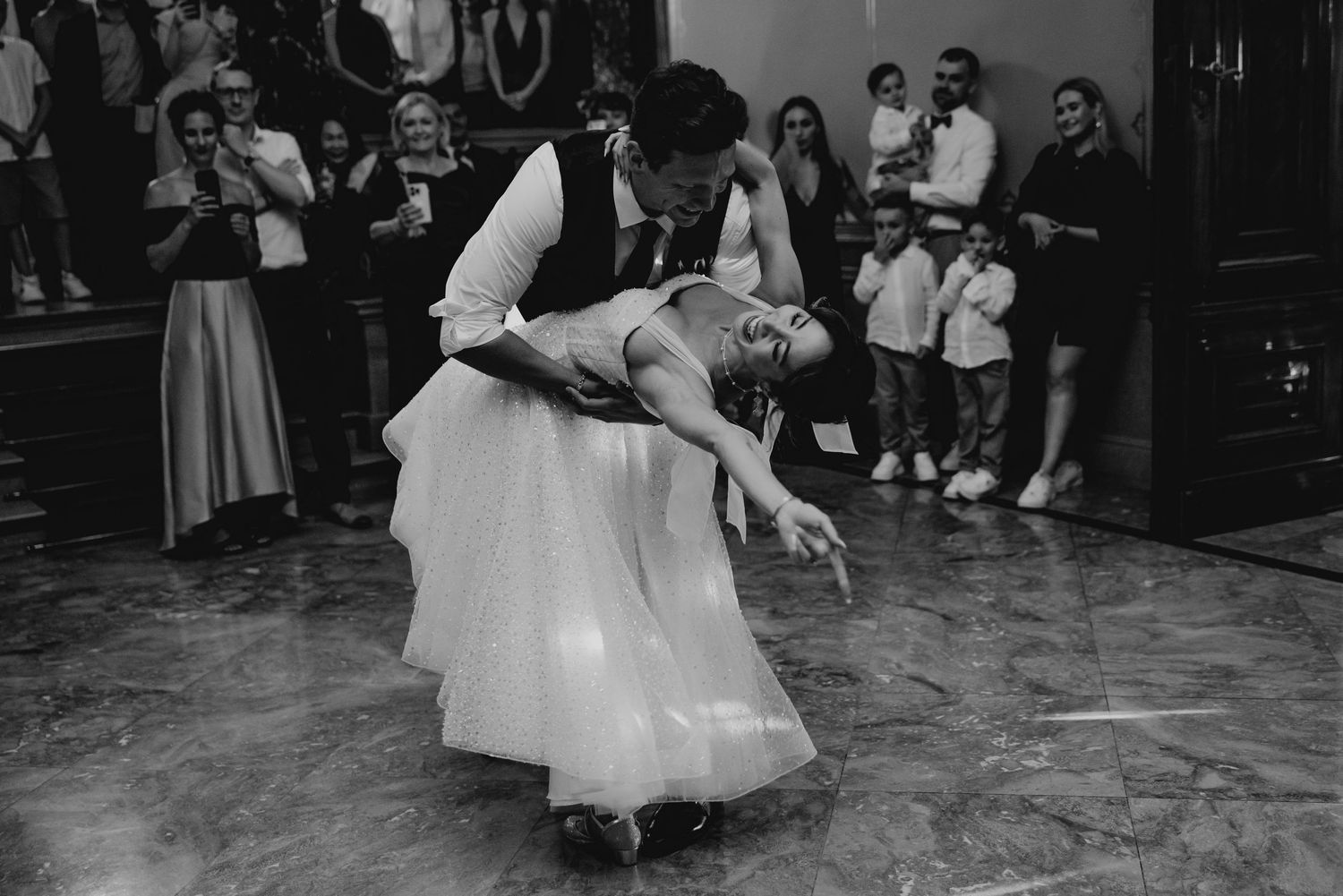 Couple performs elegant dance dip move in black and white during romantic wedding celebration with spectators watching.