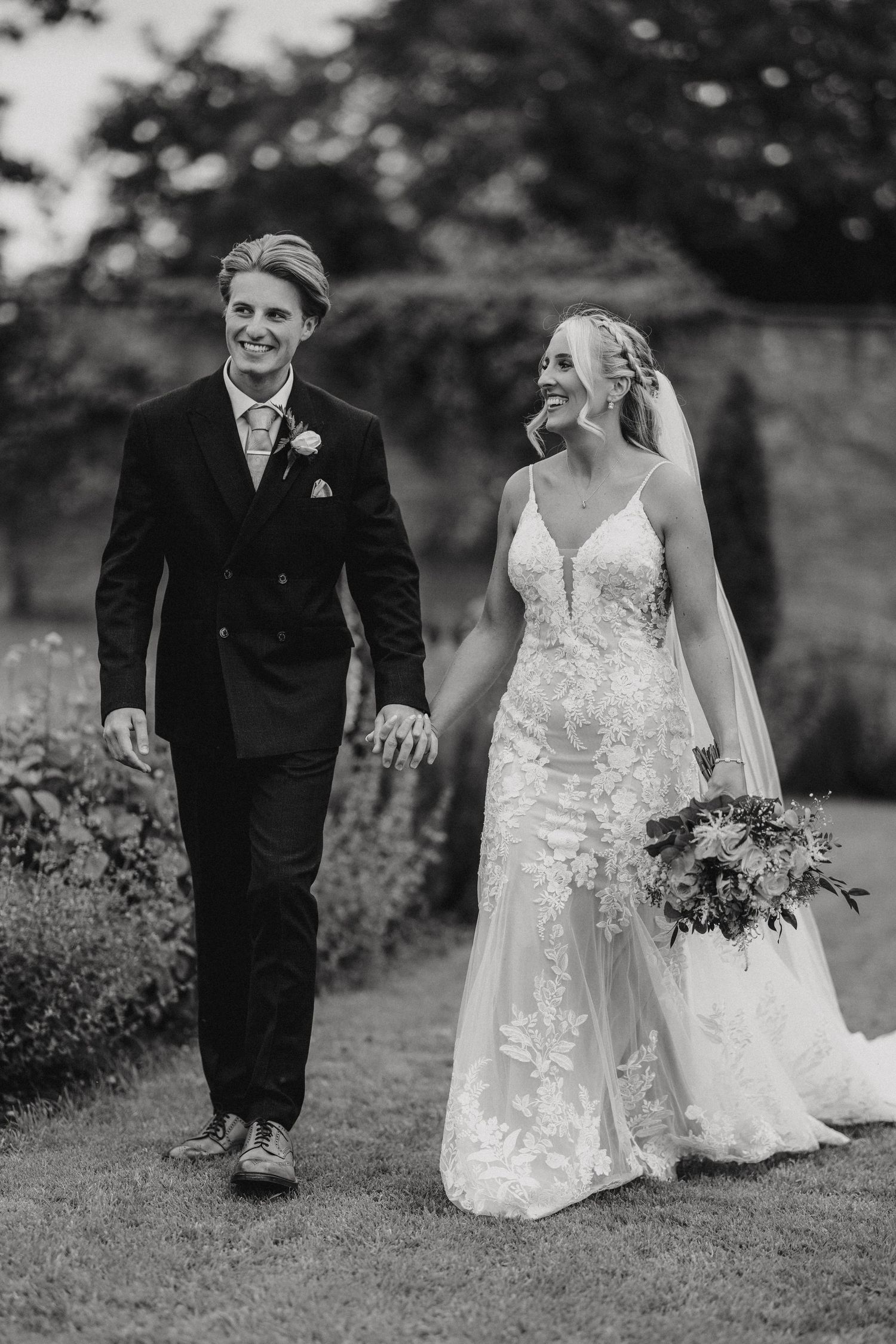 A black and white wedding photo of a couple walking together, with the bride in lace and groom in a dark suit.