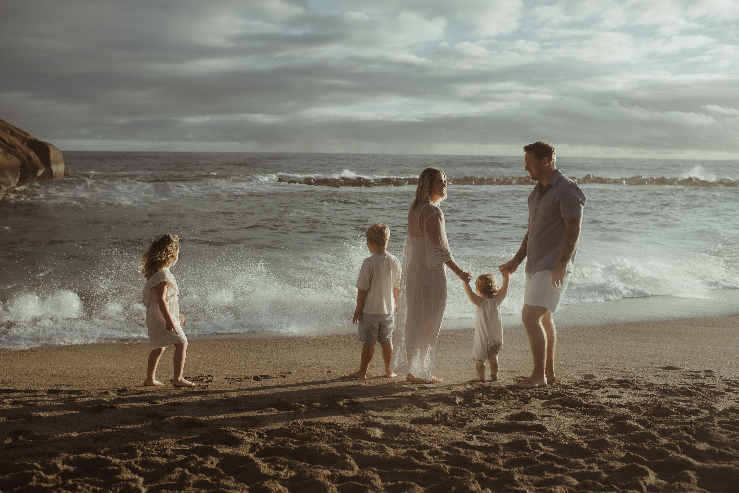 A family walks along a misty beach shoreline with dramatic stormy clouds overhead and crashing waves at Playa del Duque.