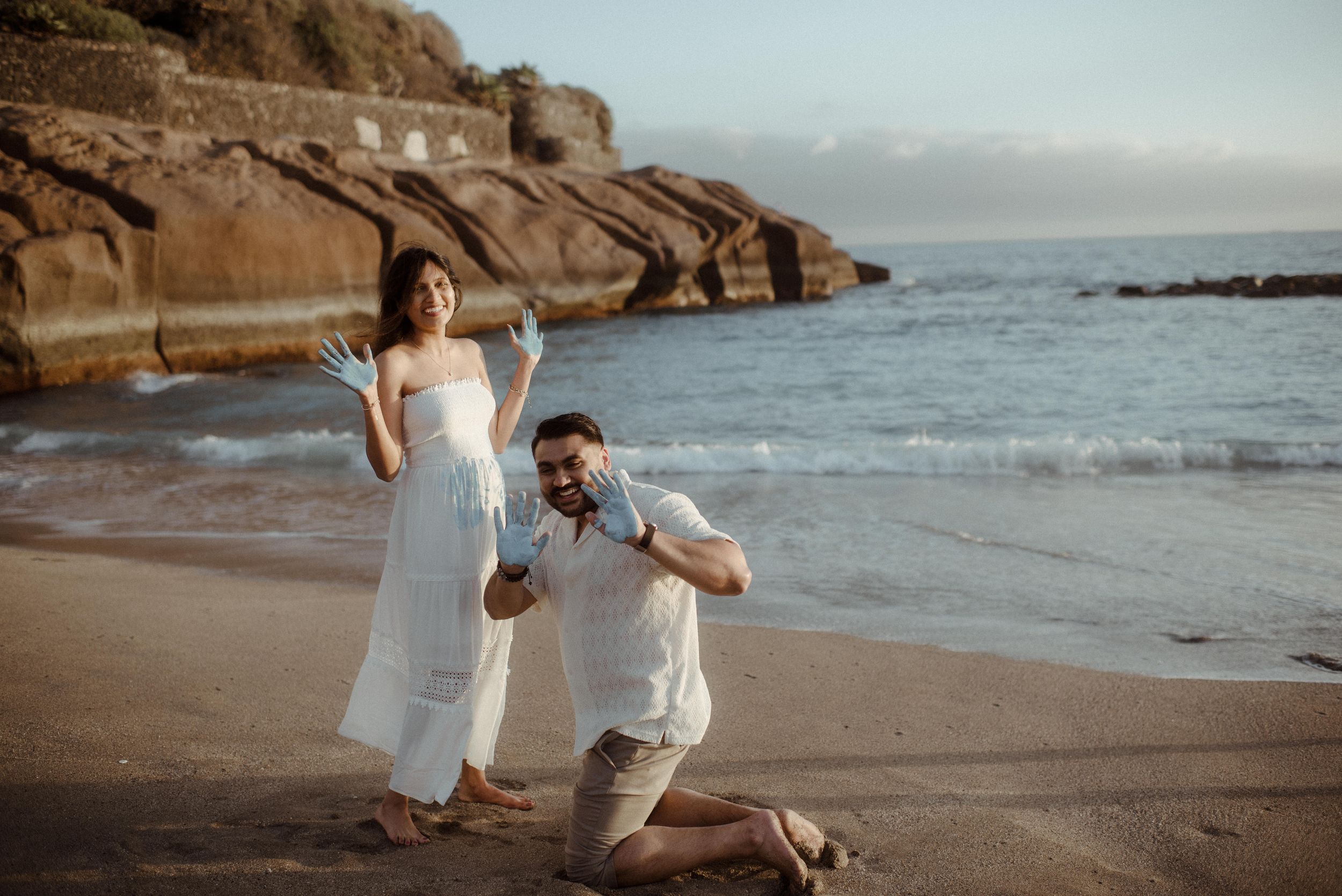 A couple in white attire poses on a scenic beach with rocky cliffs during golden hour. Gender reveal at Playa del Duque..