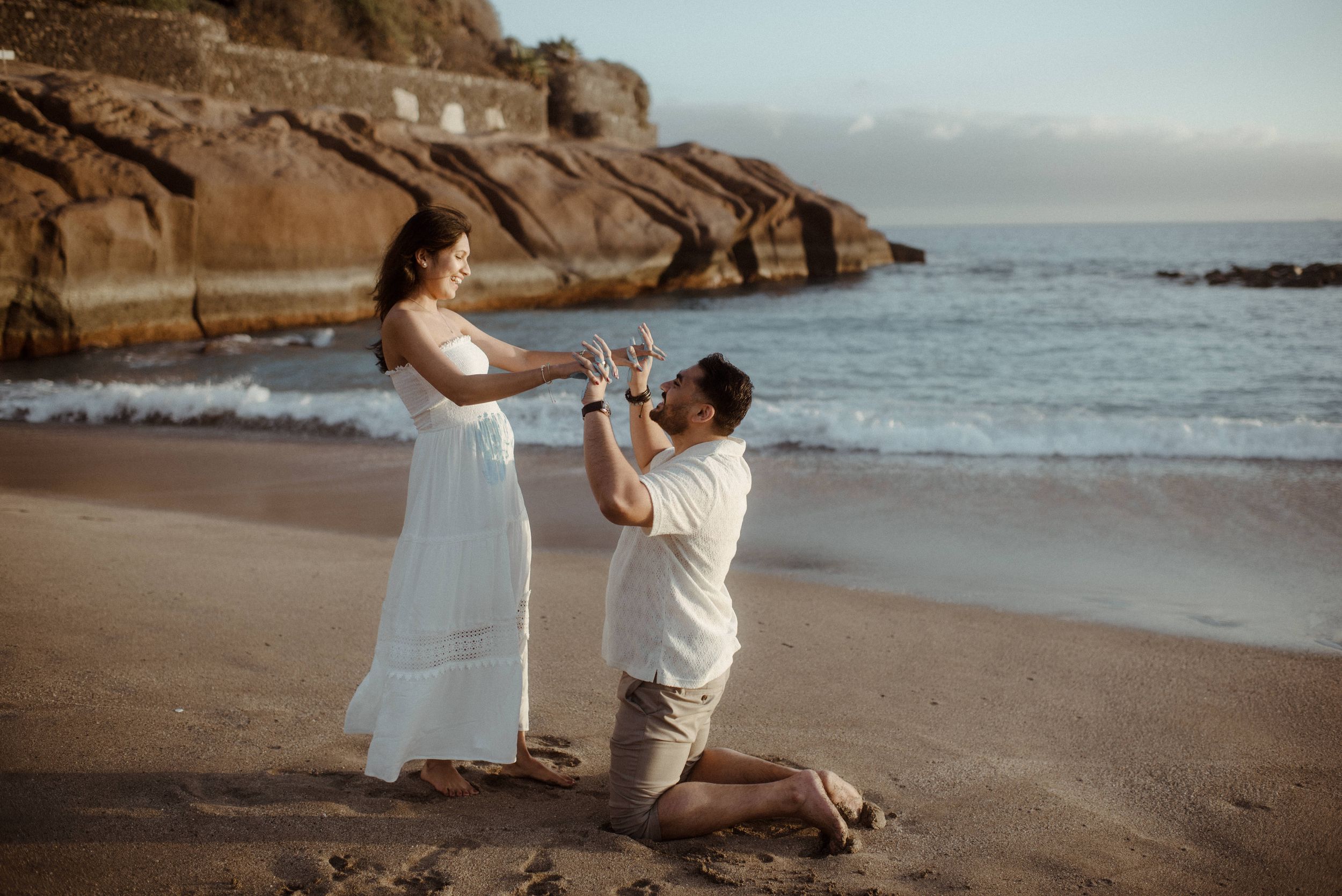 A couple shares a romantic moment at sunset on a scenic coastal beach surrounded by cliffs and ocean waves. Gender reveal at Playa del Duque