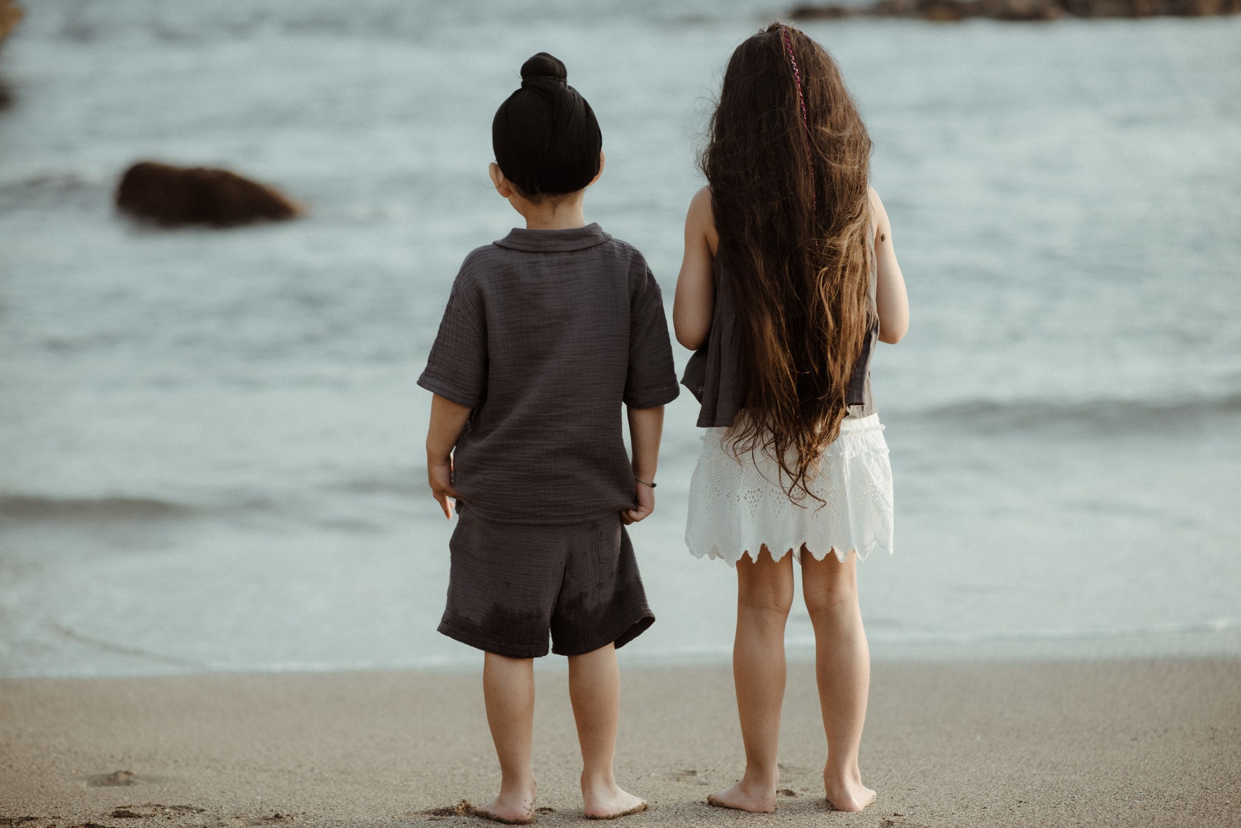 Two children stand at water's edge on a sandy beach watching waves roll in at sunset at Playa del Duque.