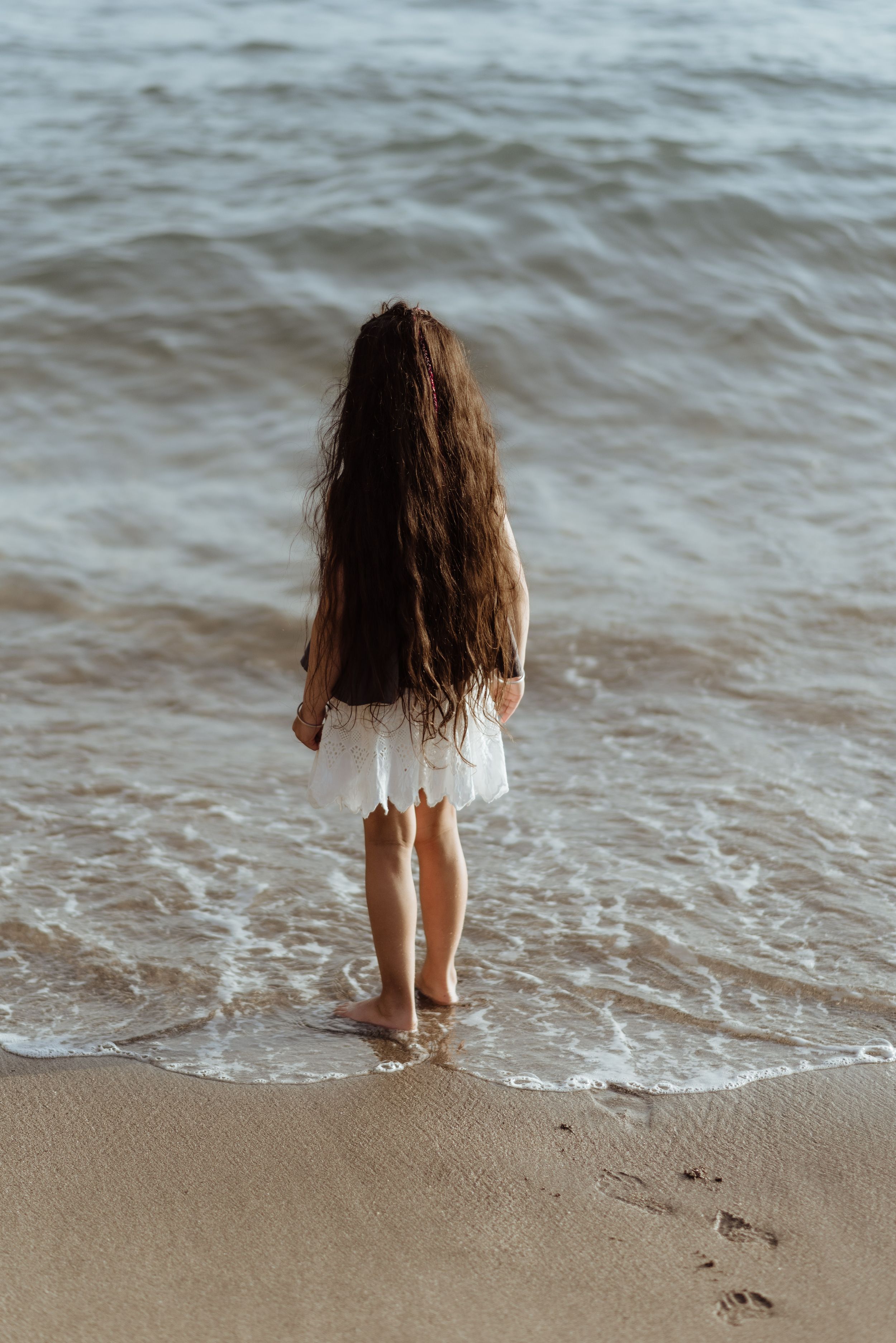 A girl in a white dress with long dark hair stands at the edge of the ocean waves on a sandy beach at Playa del Duque, Tenerife.