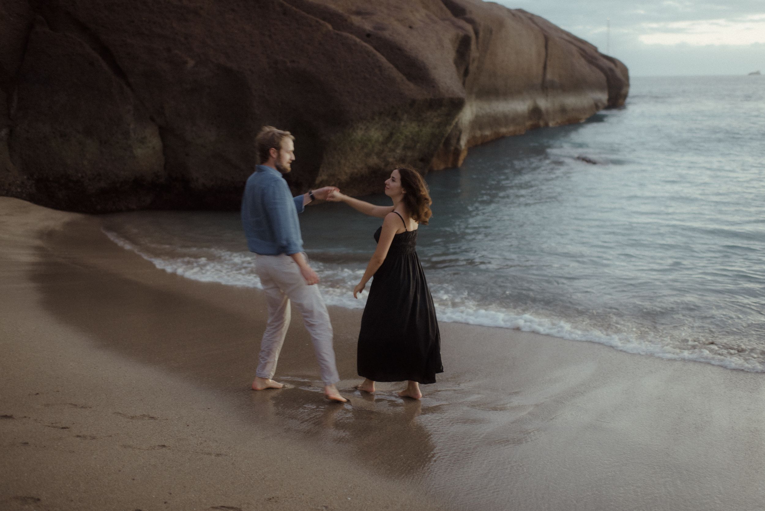 A couple walks along a scenic beach at sunset with rocky cliffs in the background.
