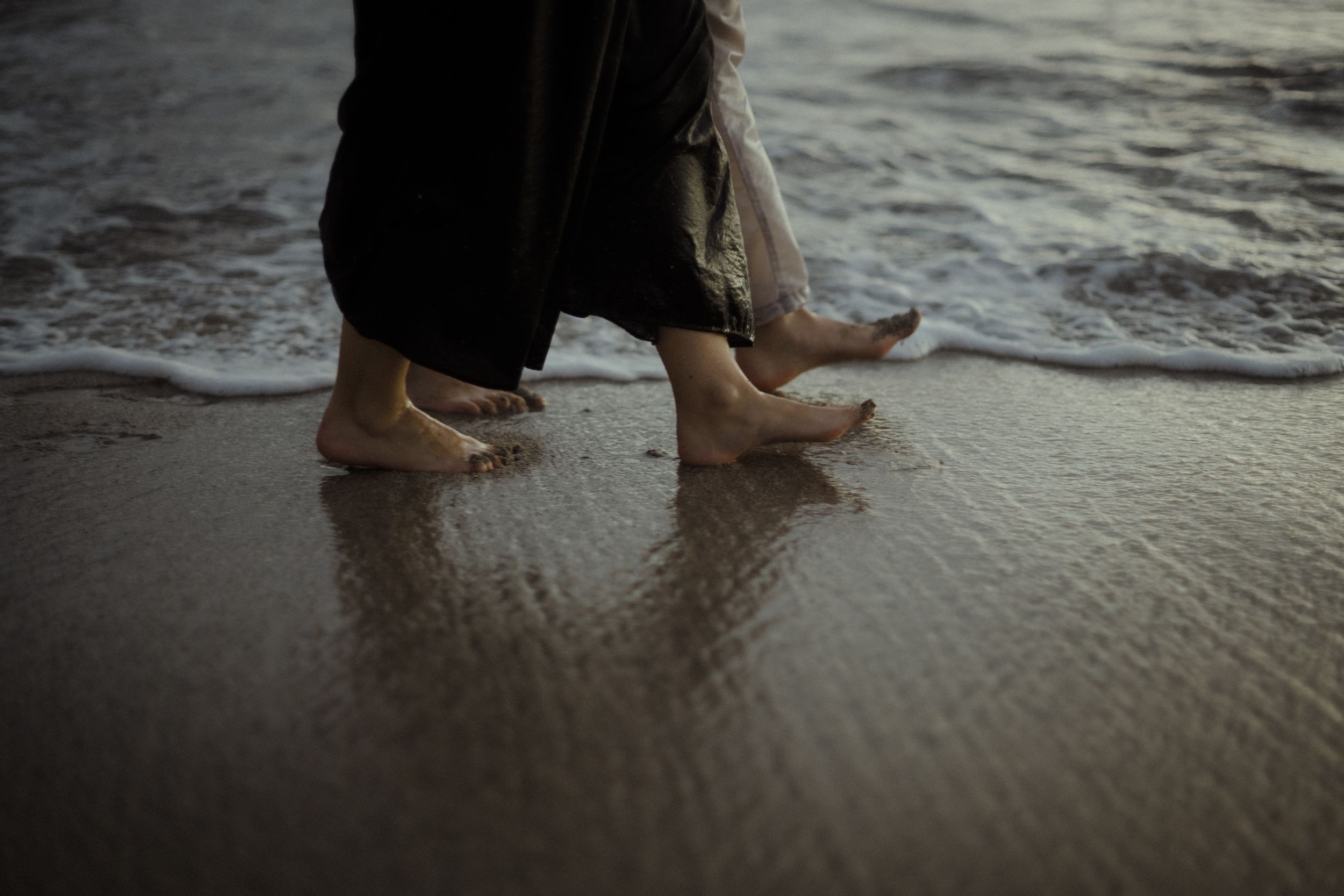 Feet walking along the water's edge on a sandy beach at dusk.