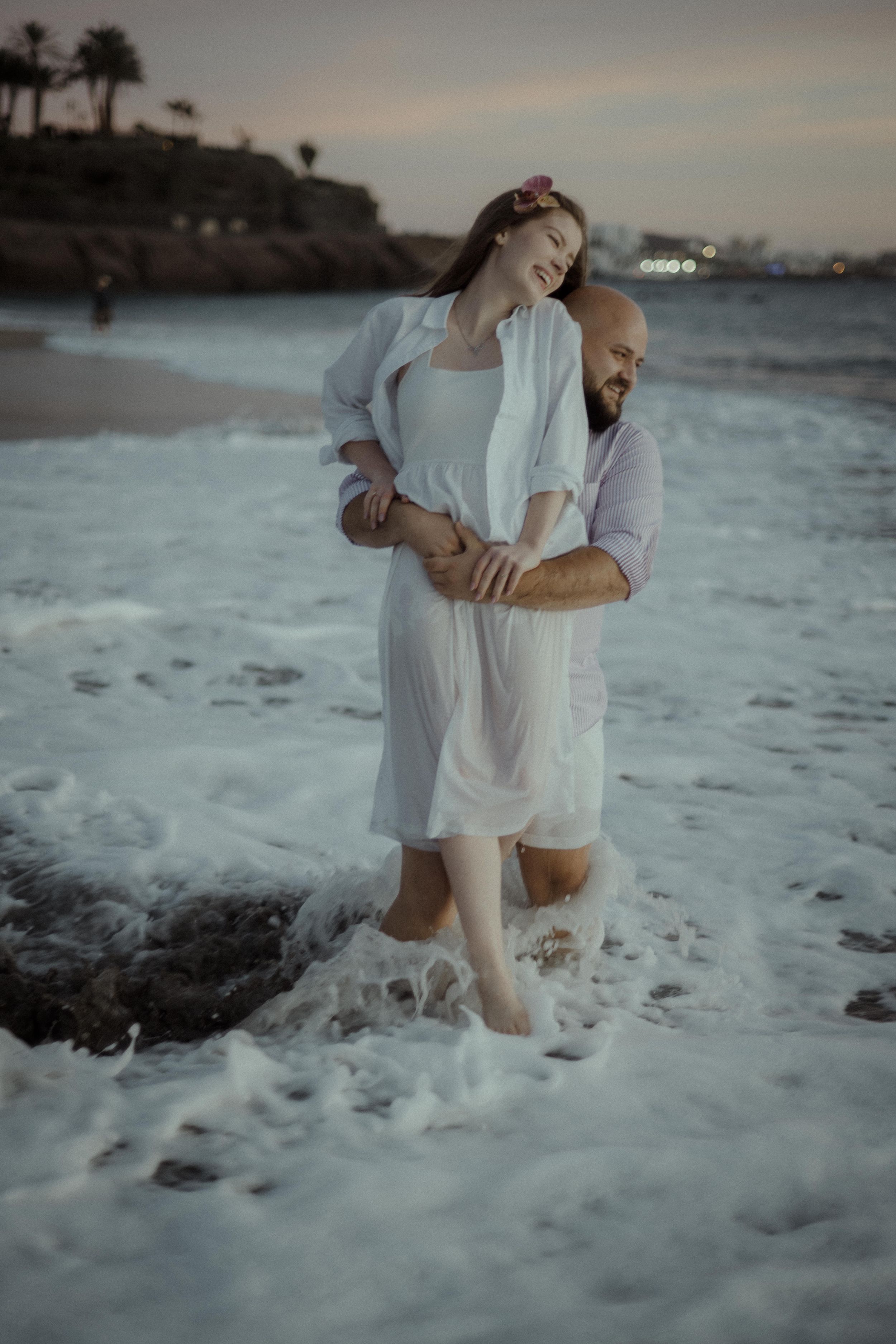 A couple embraces while standing in ocean waves at sunset on a beach with palm trees in the background.