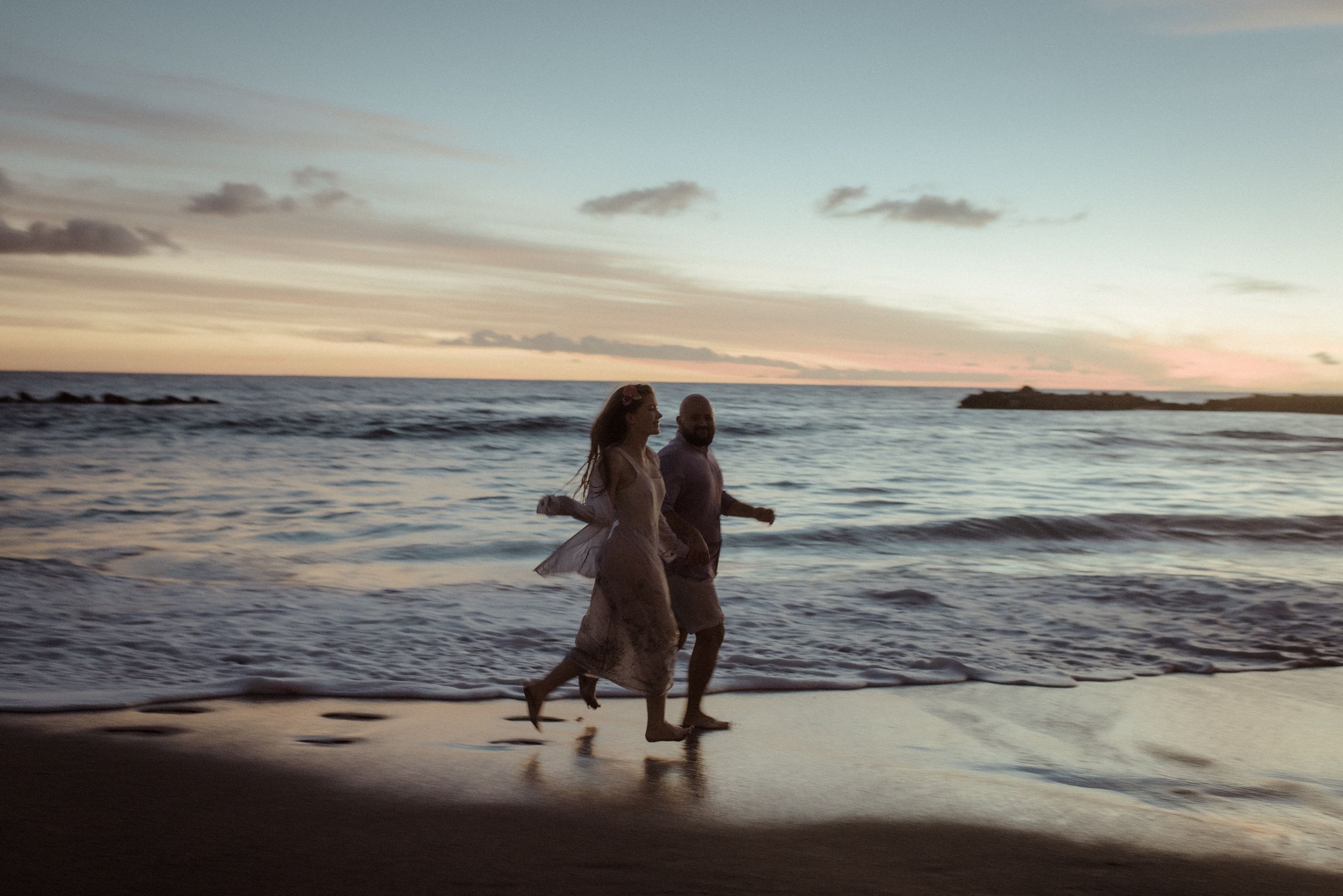 Silhouetted figures walk along a beach shoreline at sunset with waves crashing against the sand.
