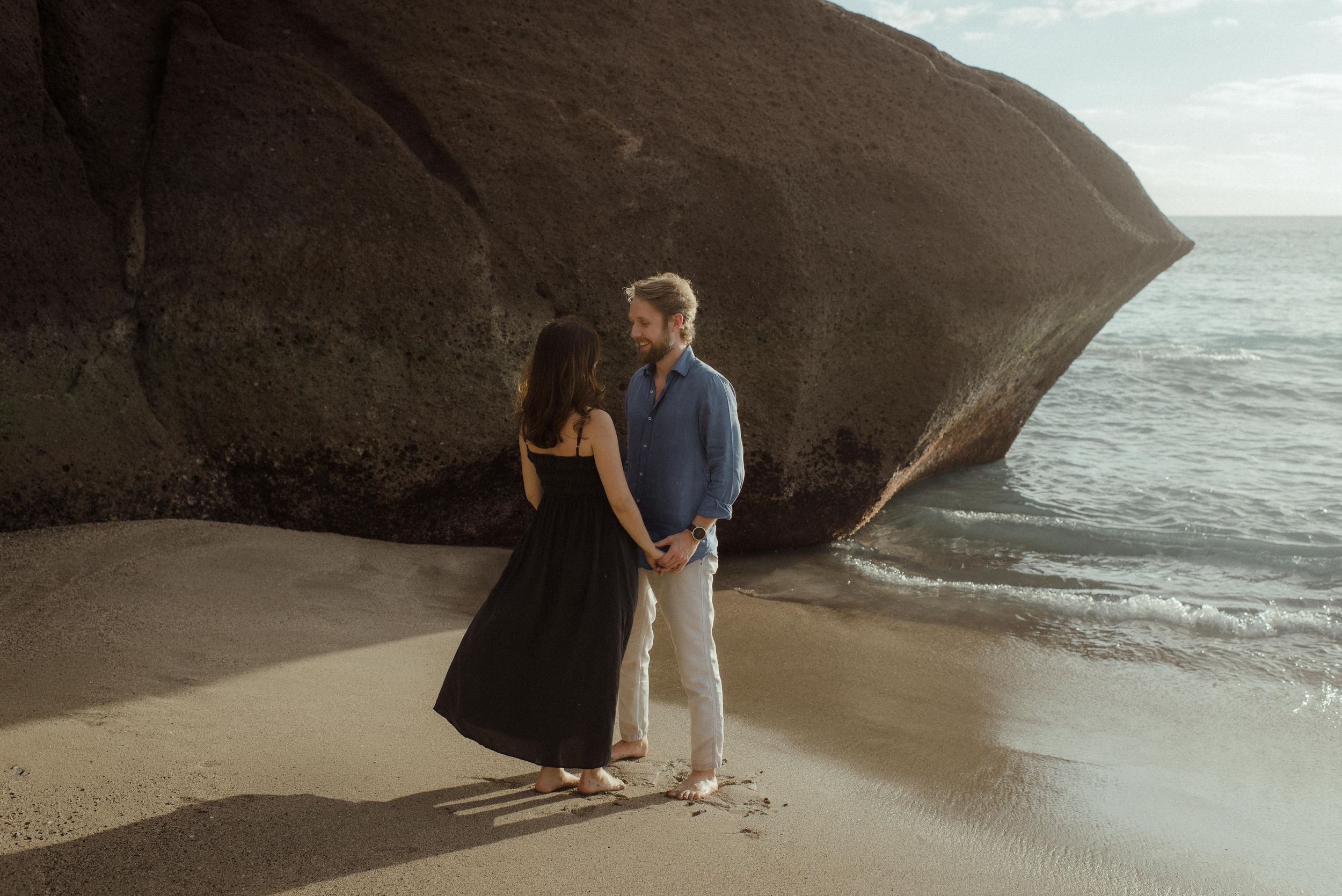 A couple stands together on a sandy beach at sunset with dramatic coastal rock formations in the background.