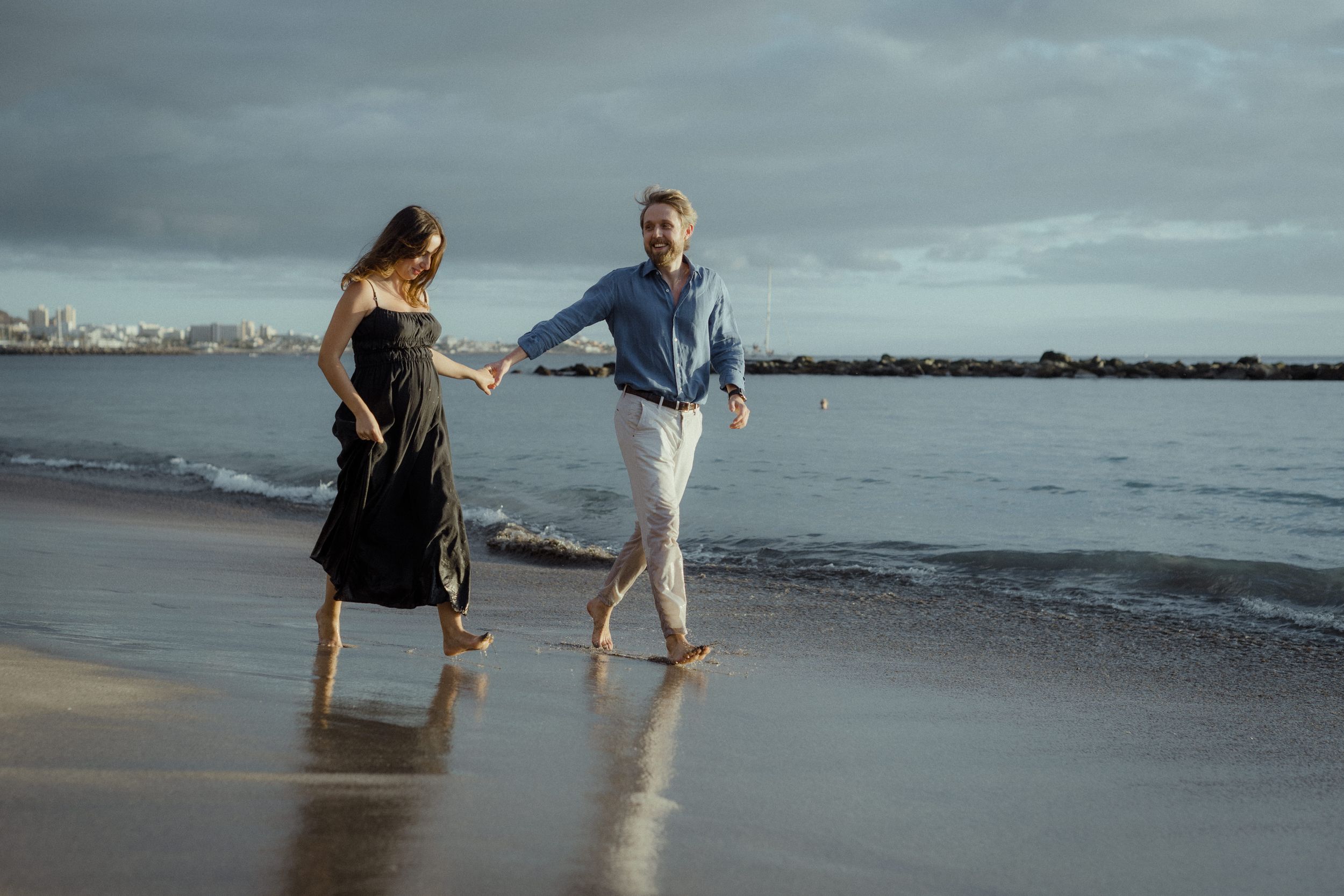 Two people holding hands while walking barefoot along a moody beach at sunset with city skyline in background.
