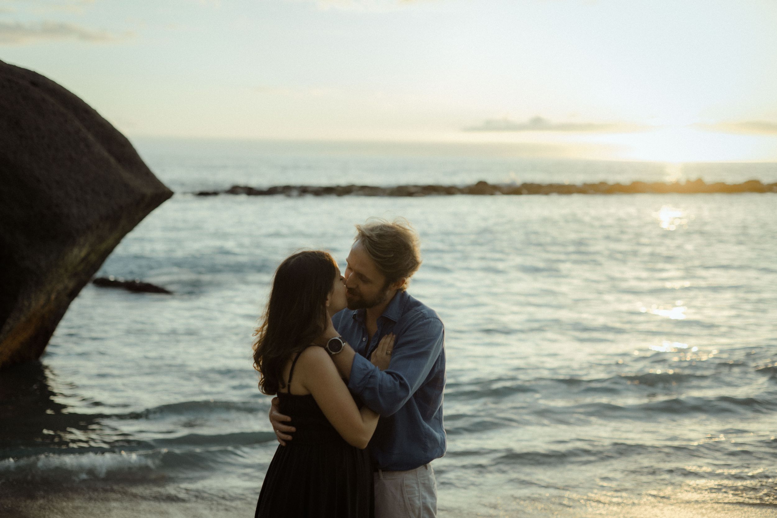 Romantic couple embraces on beach during golden sunset with ocean waves and rocky coastline in background.