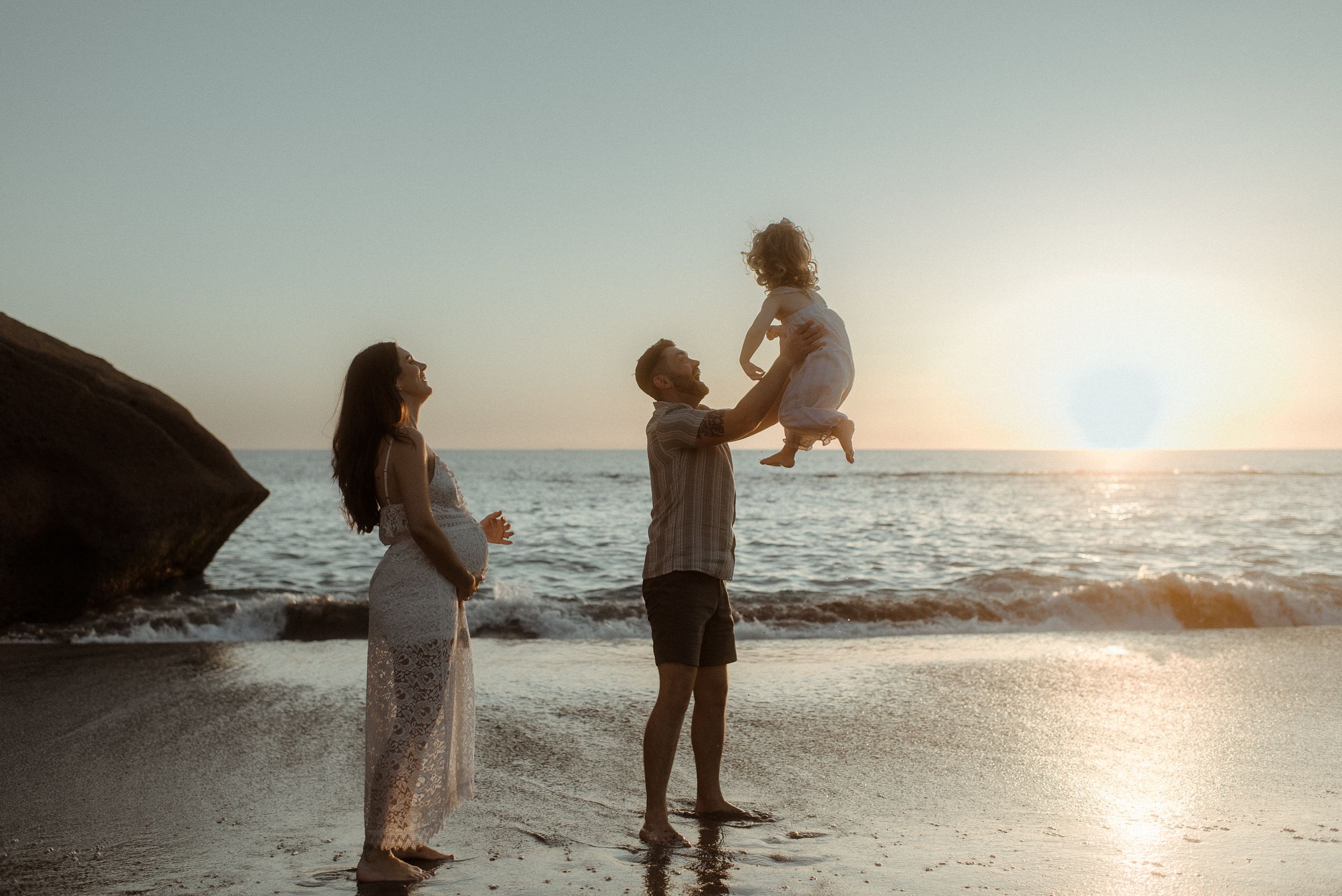 A family enjoys a tender moment together at sunset on a beach with ocean waves and rocky cliffs in the background at Playa del Duque.