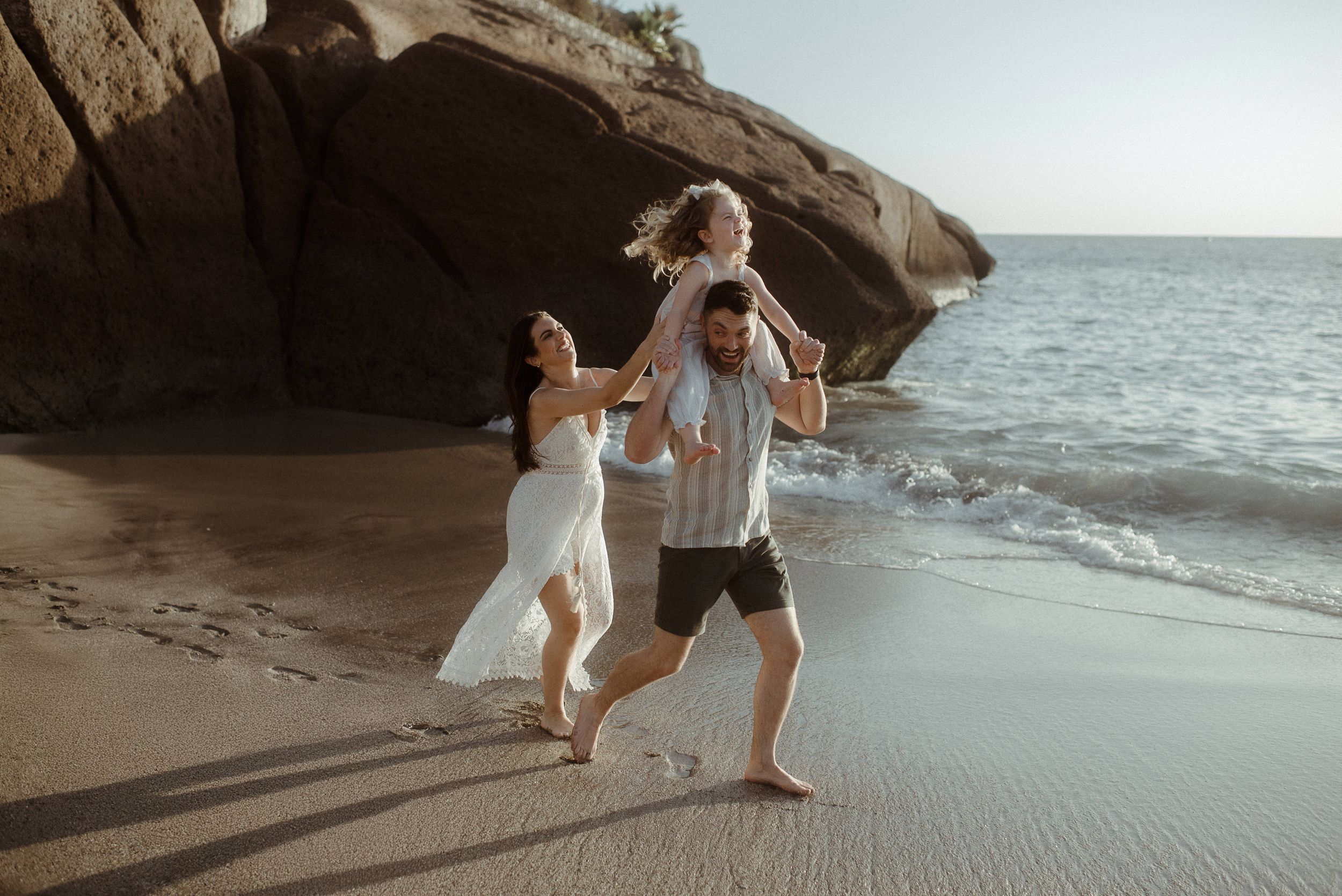 Two people in white clothing dance joyfully together on a beach near rock formations at sunset at Playa del Duque.