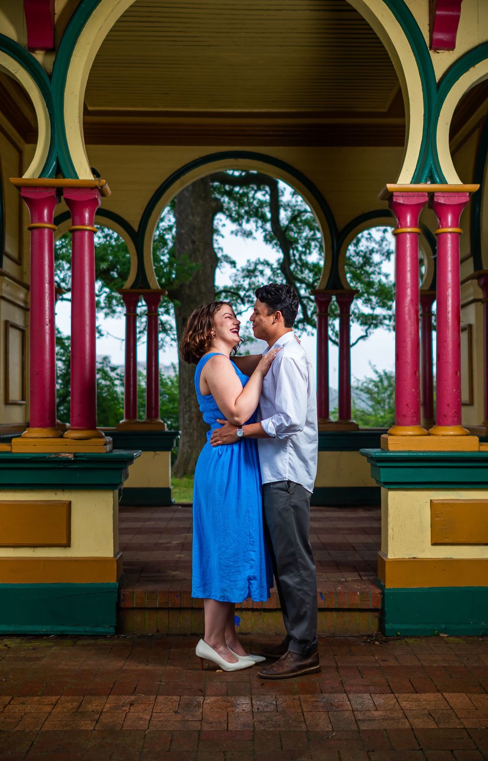 A couple shares a romantic moment under a colorful Victorian-style gazebo with pink columns and arched details, in baltimore.
