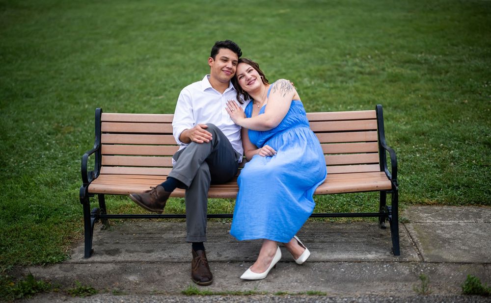 A couple relaxes together on a wooden park bench, with the person in a blue dress leaning against their partner, in baltimore