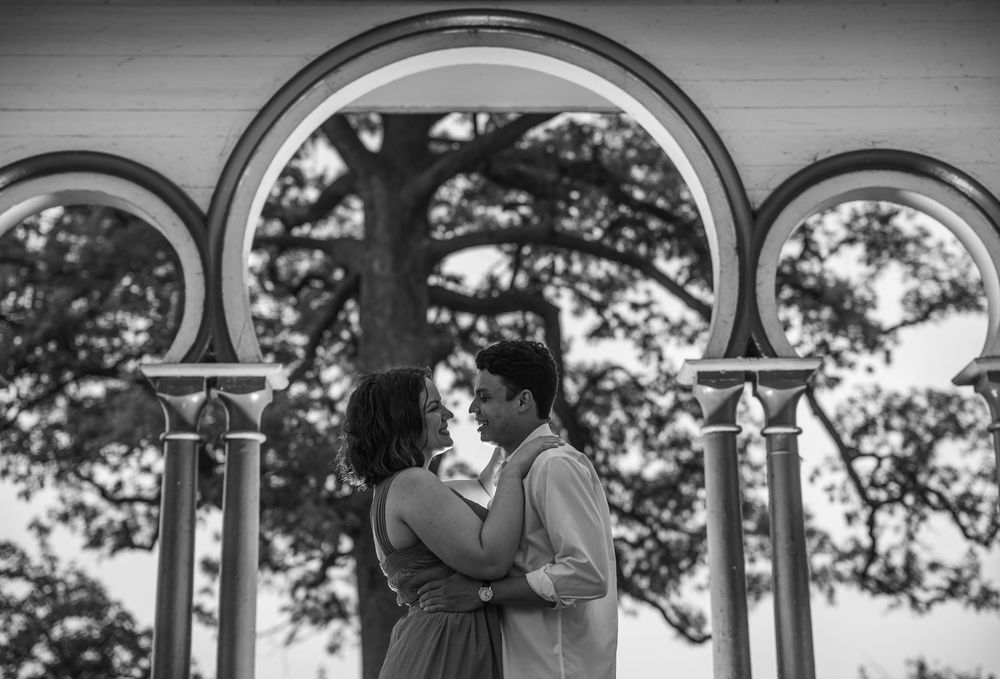 A romantic black and white portrait of a couple embracing under decorative stone arches with trees visible in the background, in baltimore