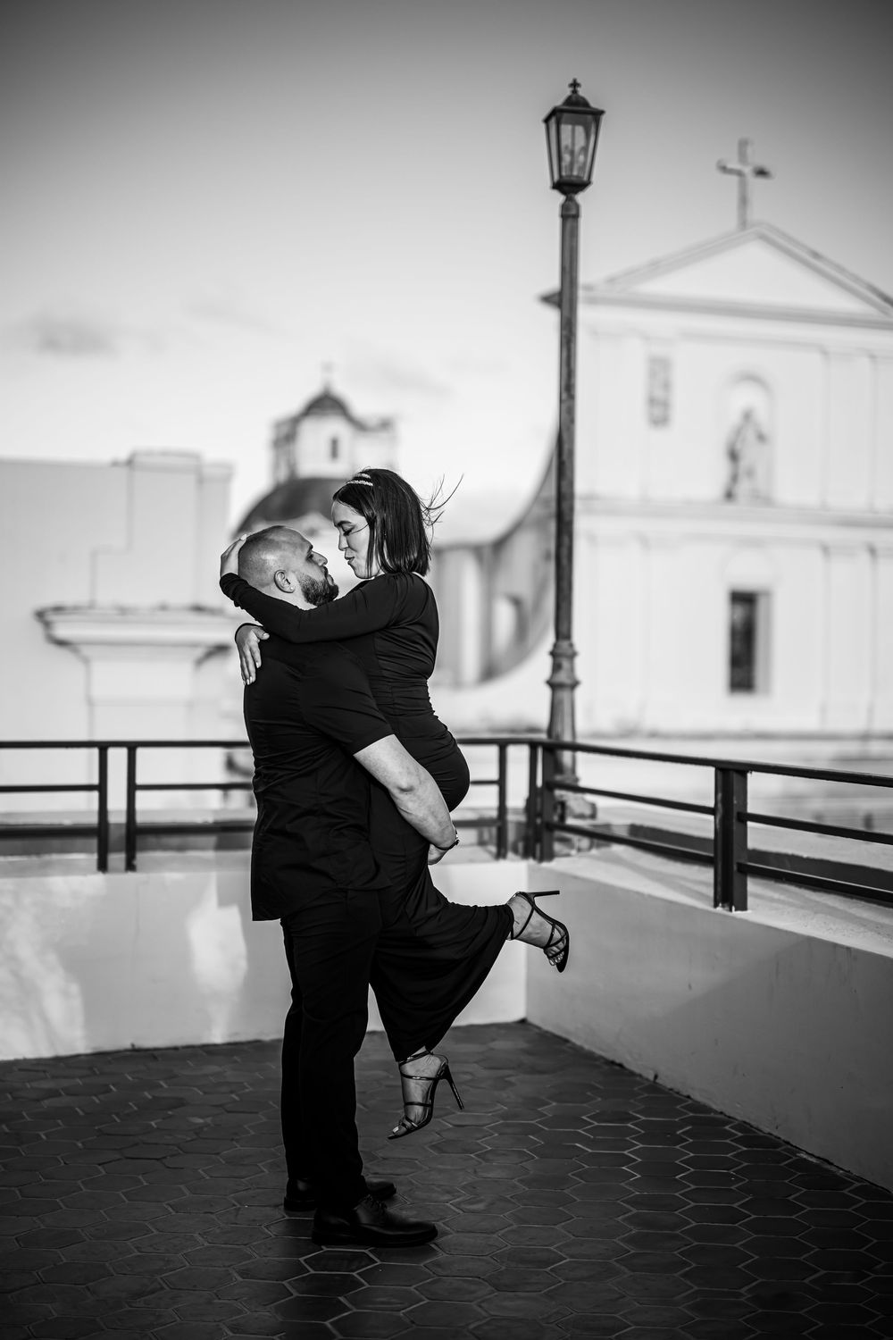 A romantic couple embracing and dancing in front of a church with a vintage lamp post in black and white photography.