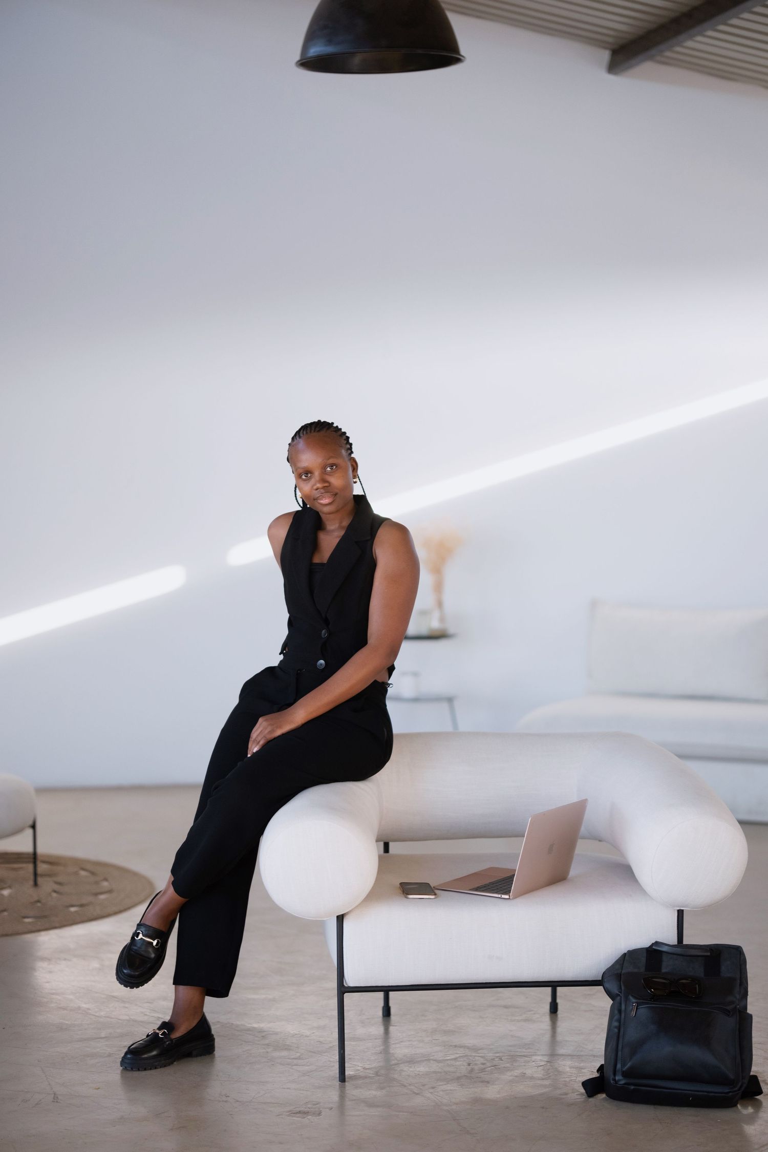 Black woman in black pantsuit sits on white sofa with laptop in bright studio.