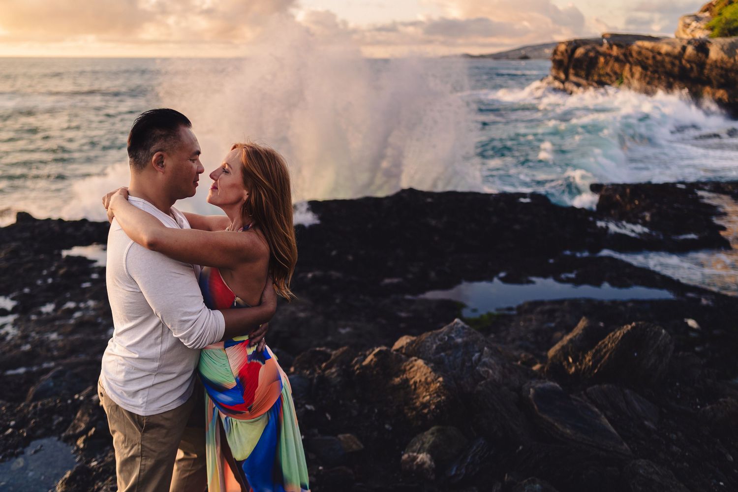 A couple embraces on rocky table rock, laguna beach coastline at sunset while ocean waves crash against volcanic rocks in the background