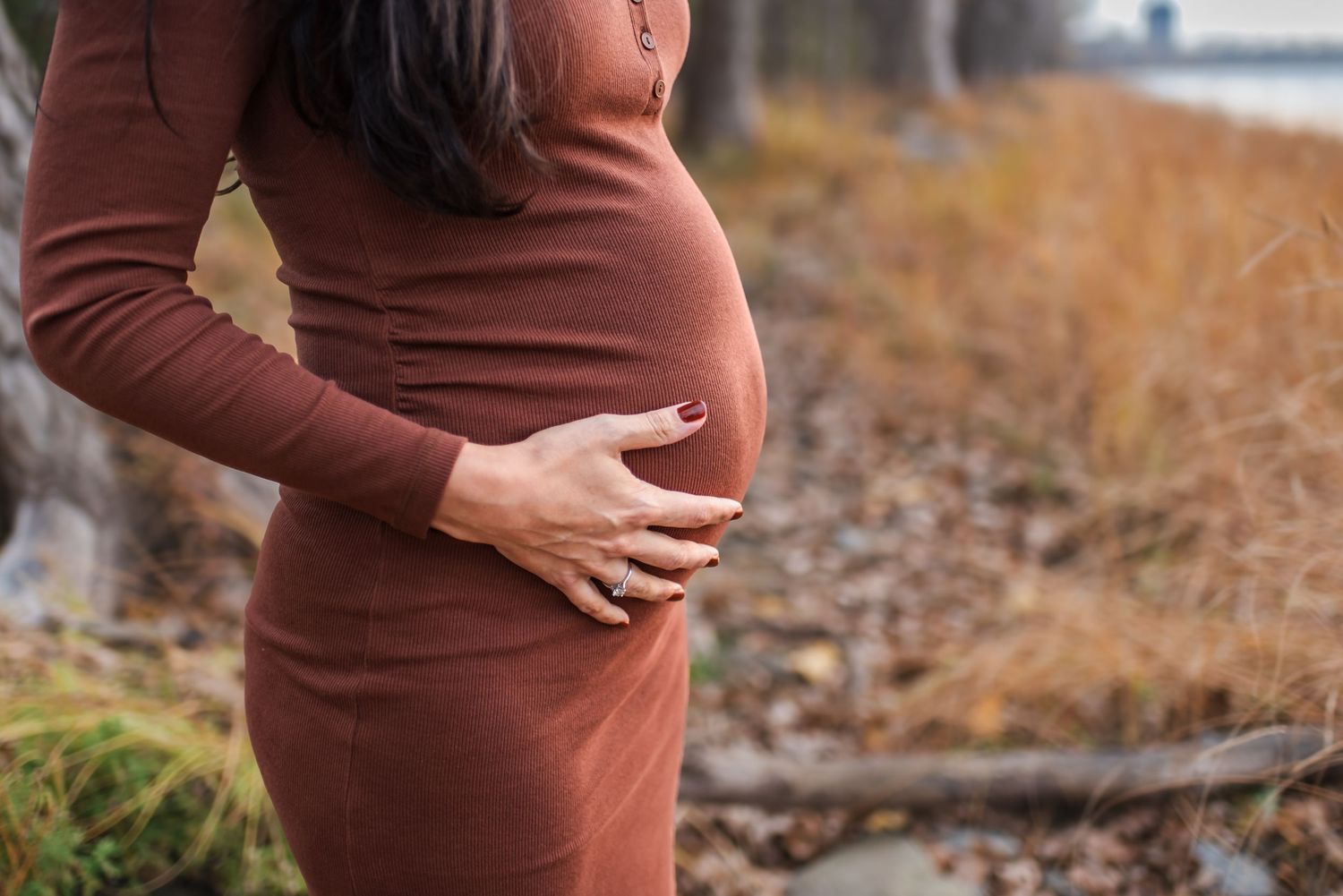 A pregnant woman in a brown sweater dress gently cradles her baby bump while standing outdoors in fall foliage.