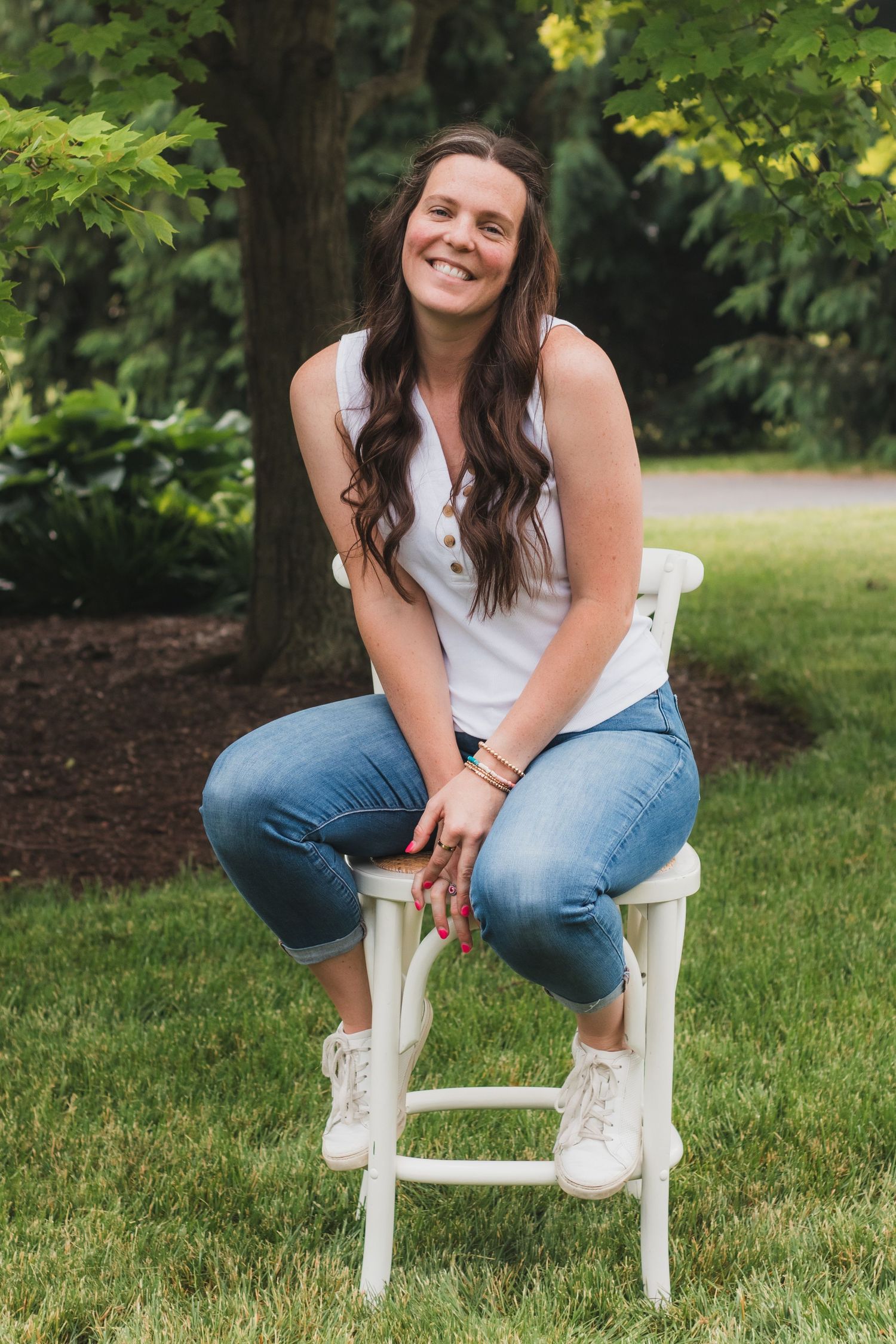 Smiling brunette woman sitting on white stool outdoors in lush green garden setting.