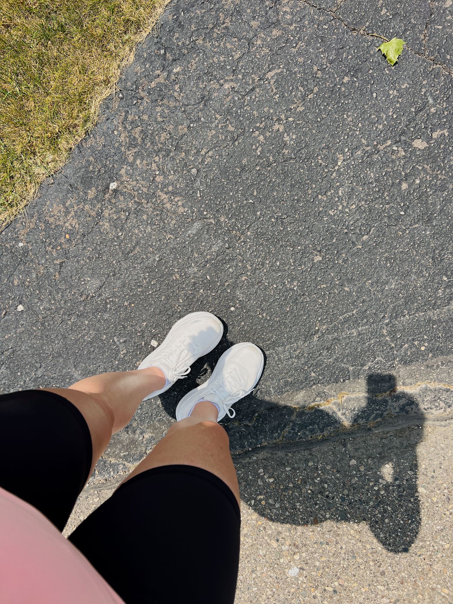 Overhead view of white sneakers on pavement with shadow visible beside green grass edge.
