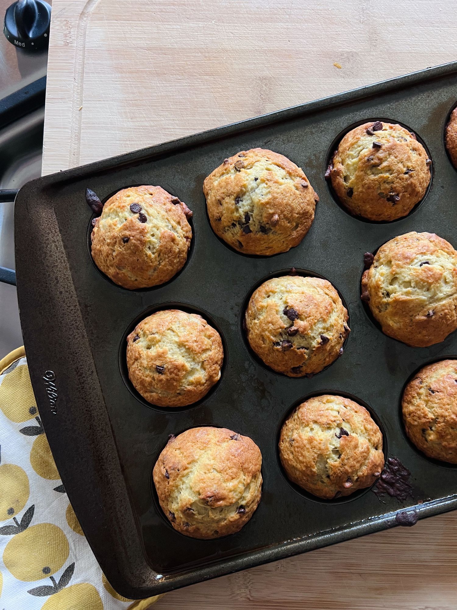 Freshly baked chocolate chip muffins cooling in dark non-stick muffin pan on wooden surface.