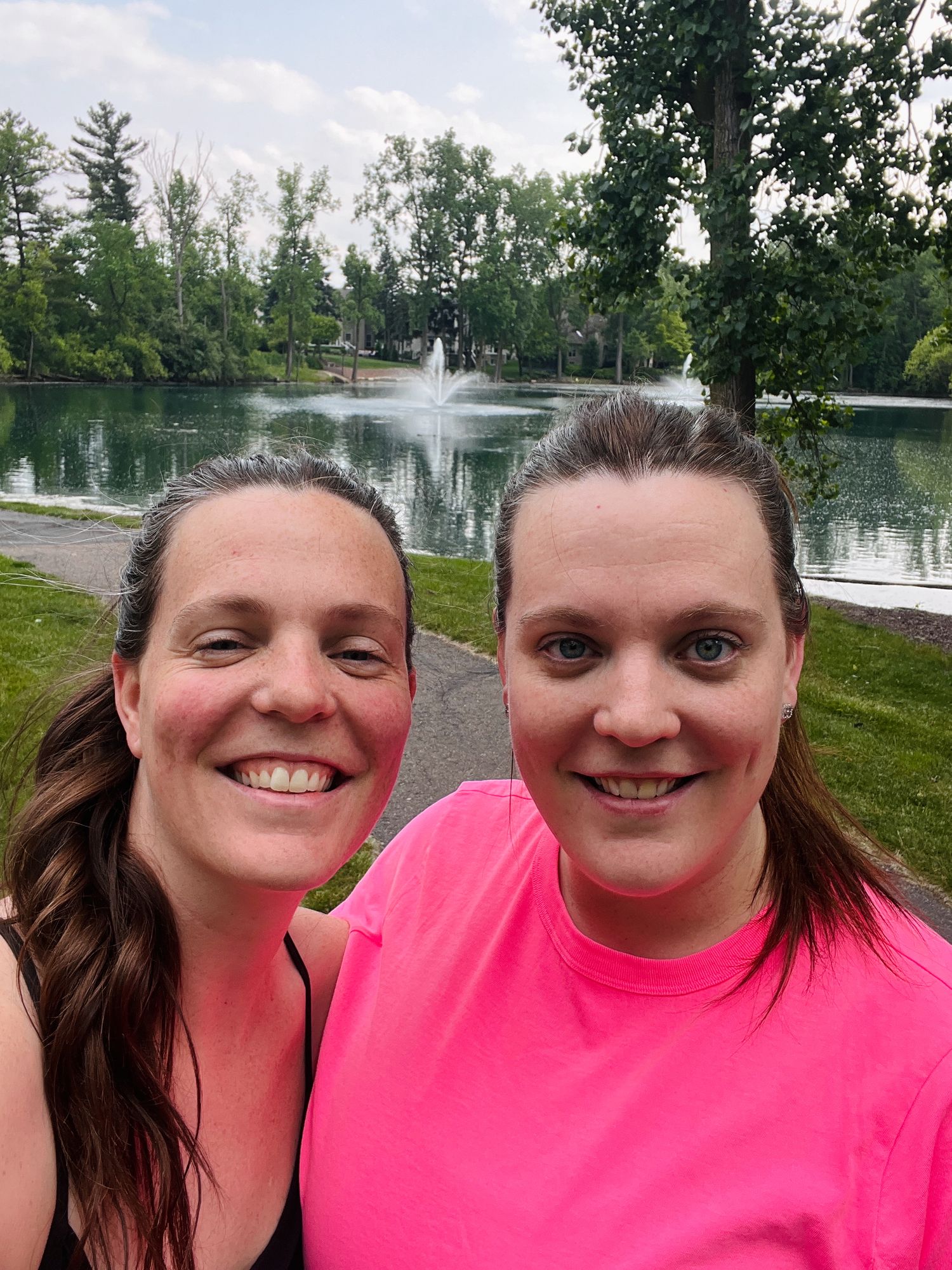Two smiling women in athletic wear taking selfie by scenic park pond with fountain.